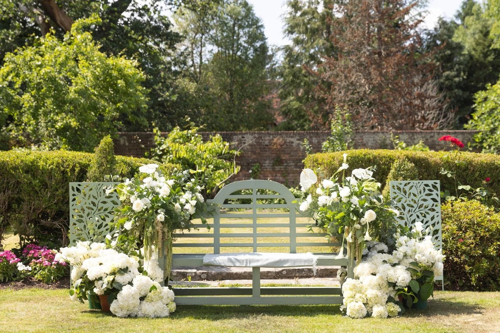 A white garden bench decorated with white flowers and lush greenery in a well-maintained garden with a brick wall and trees in the background.