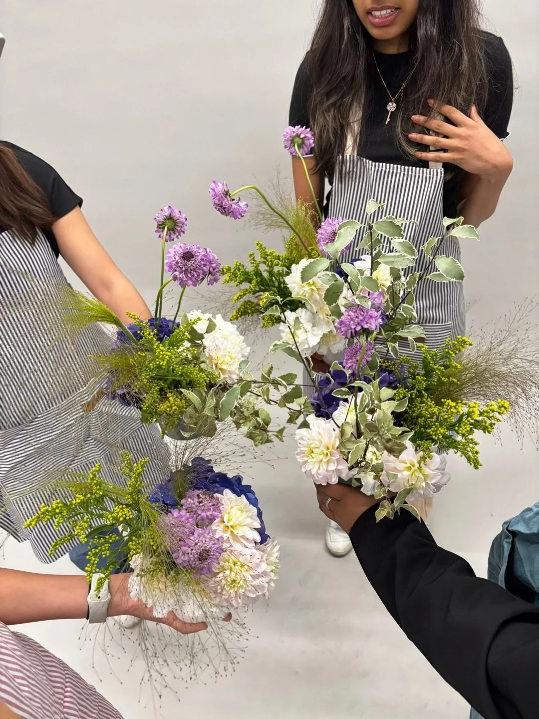 People holding vases of floral arrangements with purple, white, and green flowers.