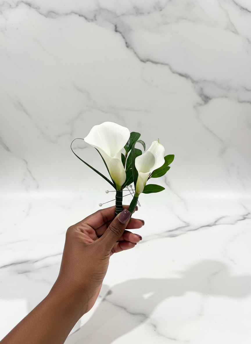 A hand holding a small artificial flower bouquet with white calla lilies and green leaves against a white marble background.