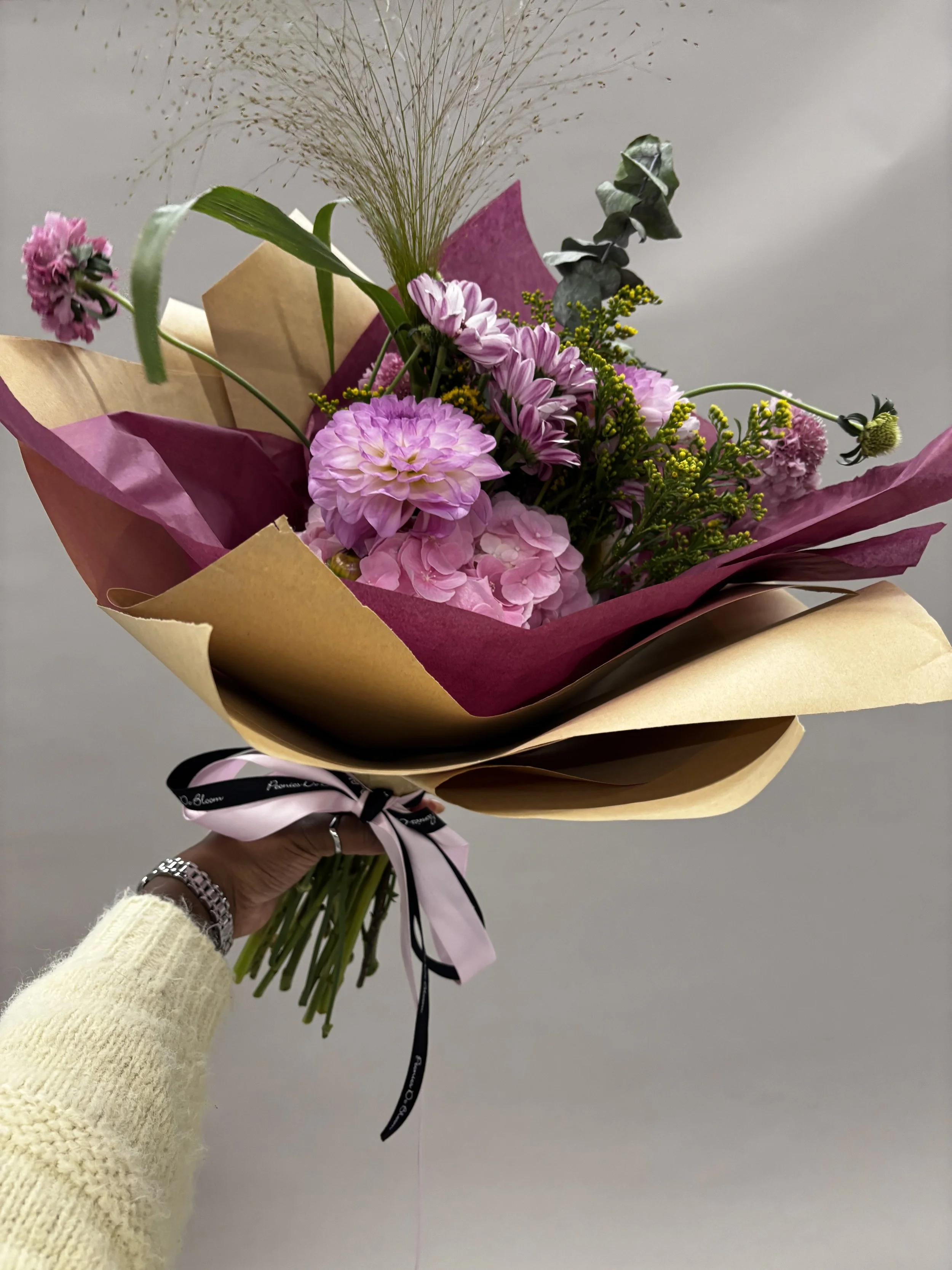 A colorful bouquet of pink and purple flowers wrapped in pink, purple, and beige paper, tied with a black and pink ribbon, held by a person wearing a yellow knit sweater.