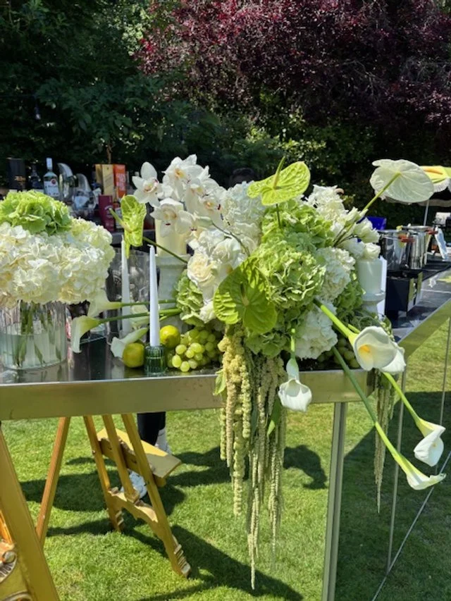 Elegant outdoor table decorated with large white floral arrangements, including calla lilies, hydrangeas, and greenery, with bottles and glasses in the background during daytime.