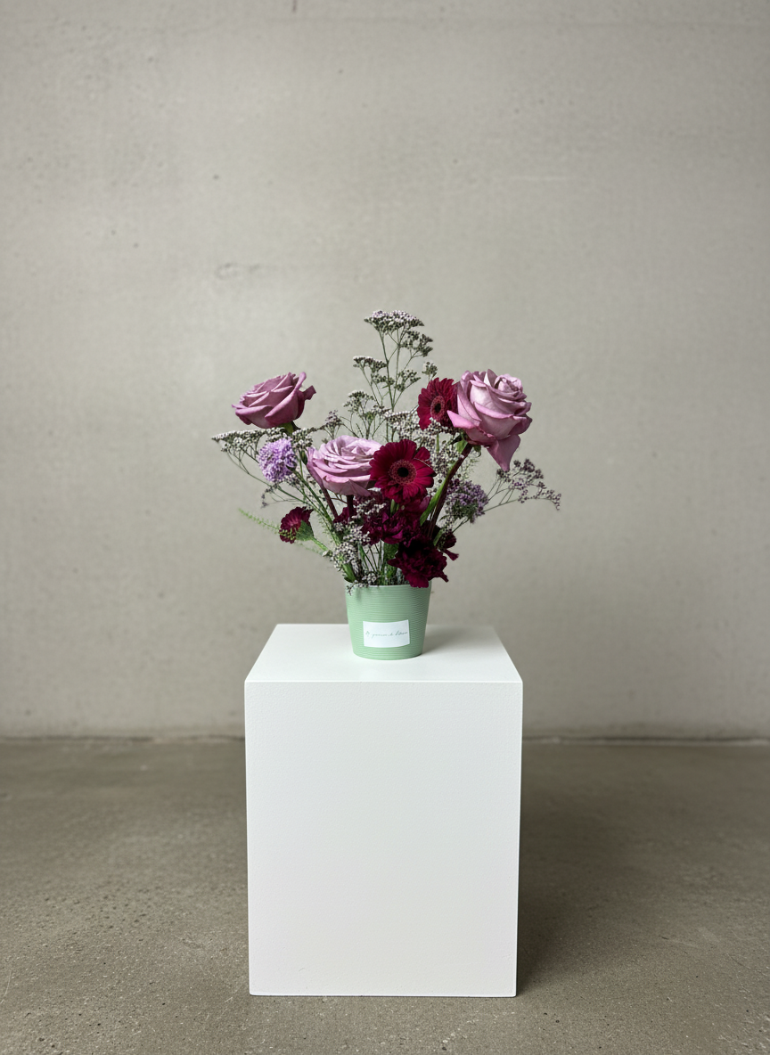 A small bouquet of pink roses, purple flowers, and red and purple blooms arranged in a pale green paper cup, placed on a white rectangular pedestal against a plain light-colored wall.