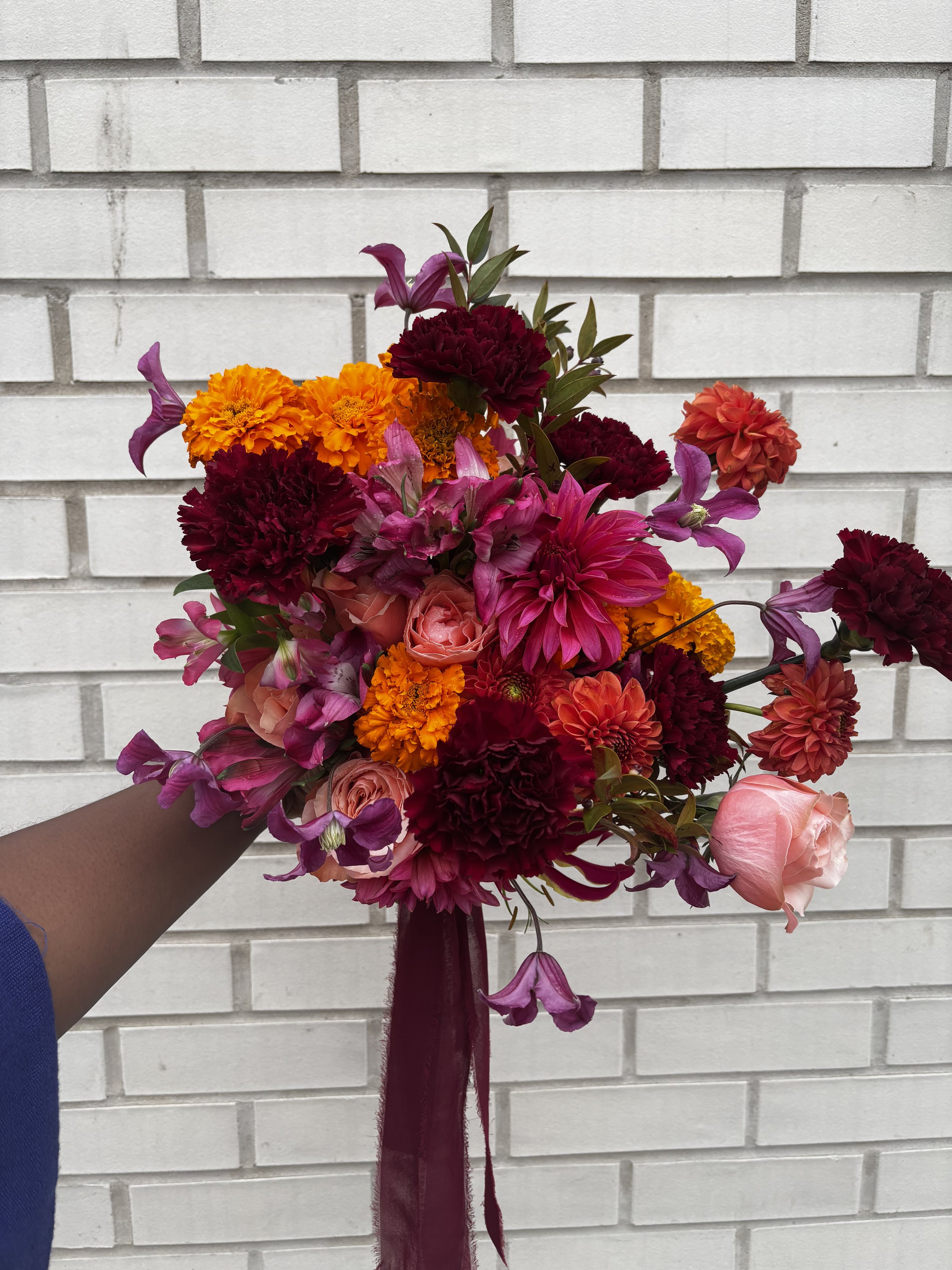 A bouquet of colorful flowers, including pink roses, orange marigolds, purple snapdragons, and deep red carnations, with a purple ribbon, held against a white brick wall background.