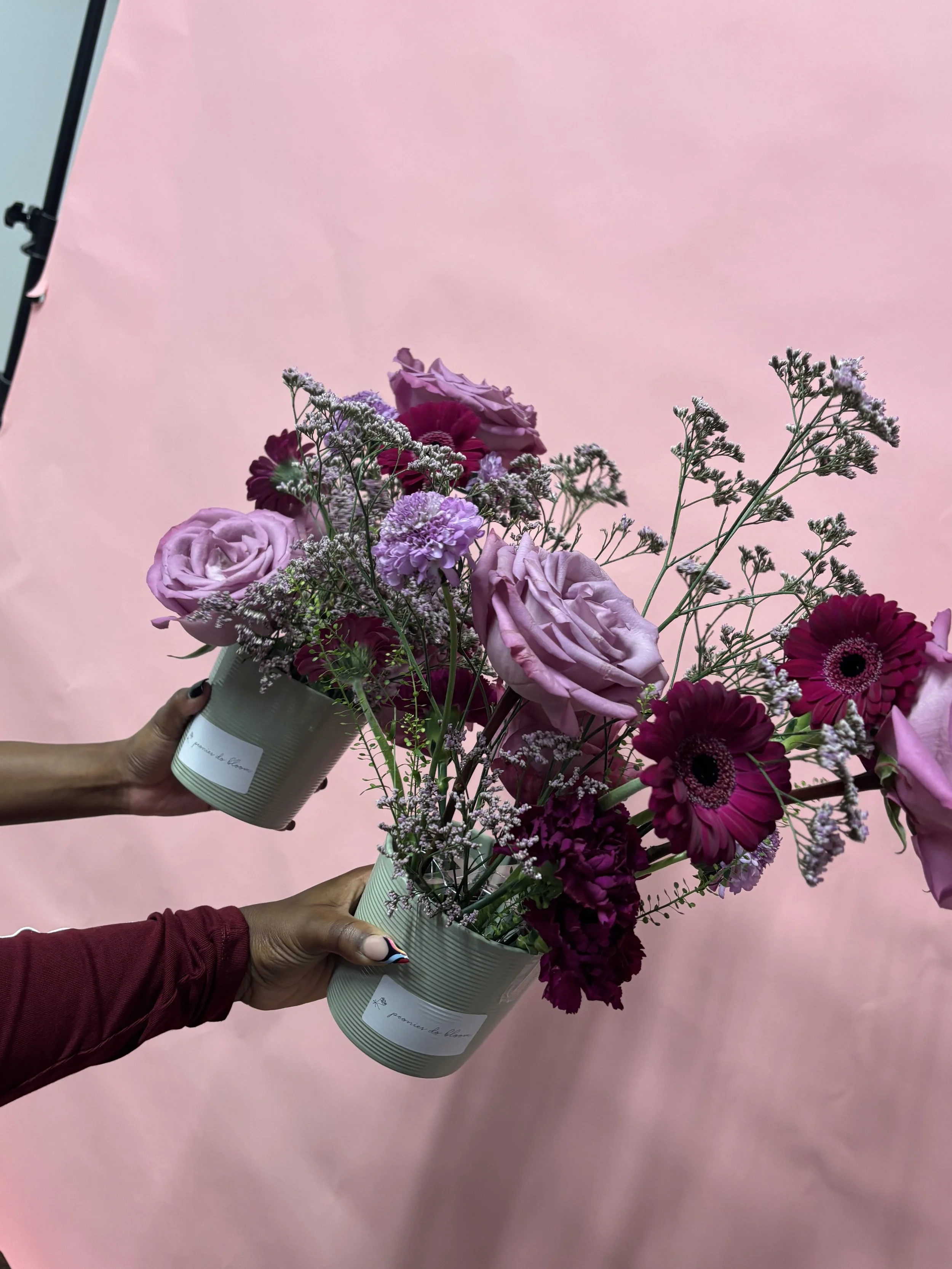 Two people holding pink and purple flower arrangements in gray pots with a pink background.