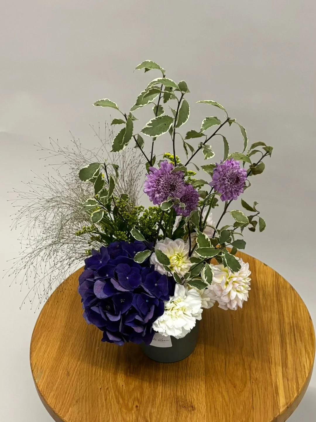 Arrangement of purple, white, and pale pink flowers with variegated green and white leaves in a black vase on a round wooden table.