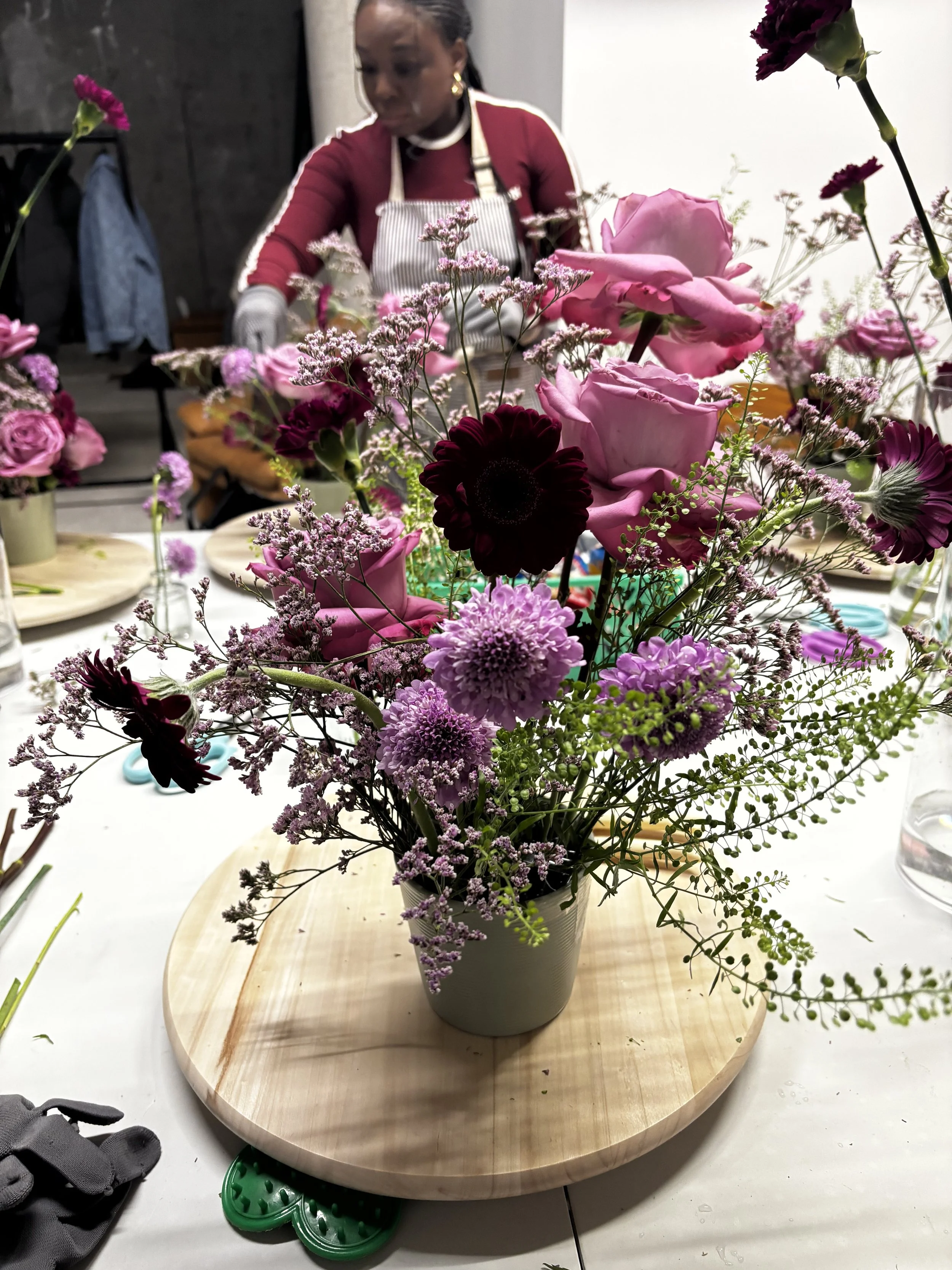 A colorful flower arrangement with pink roses, purple dahlias, dark burgundy ranunculus, and white filler flowers in a gray vase on a round wooden tray, with a woman working on floral arrangements in the background.