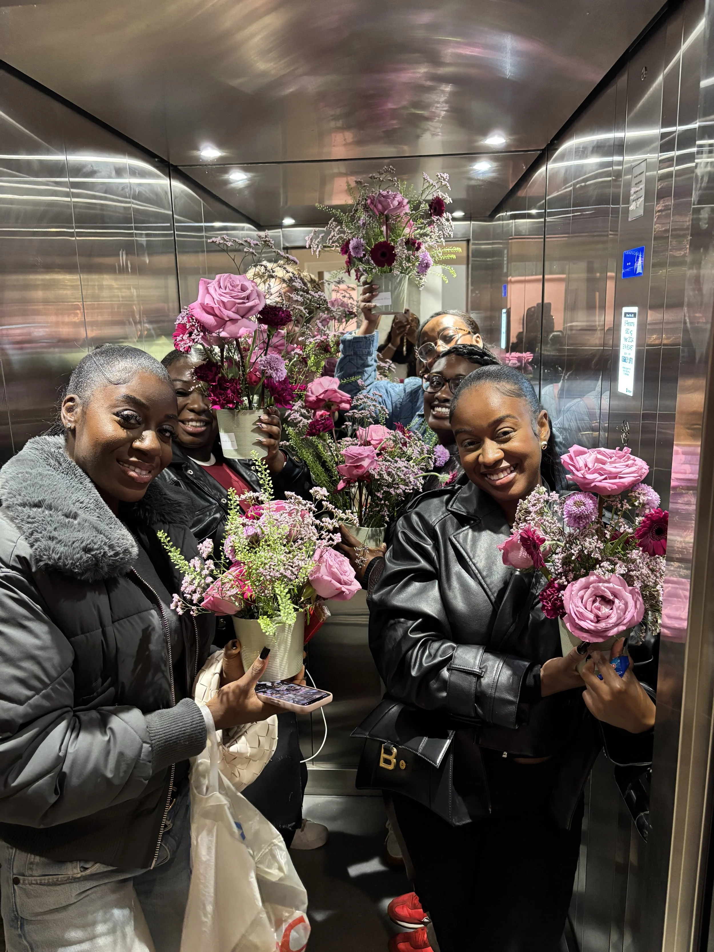 Four women in an elevator holding pink and purple flower arrangements, smiling and posing for a photo.