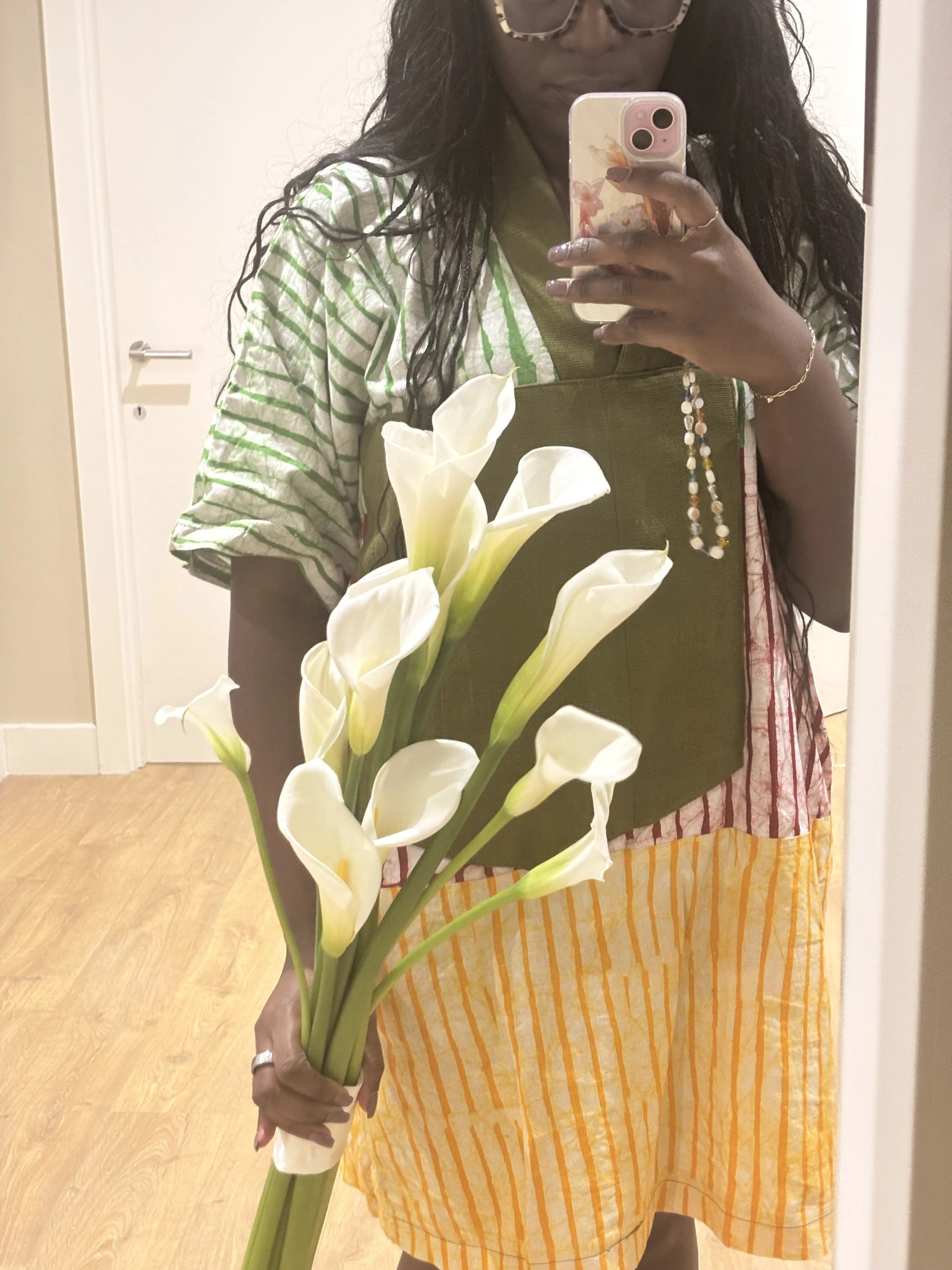 A woman taking a mirror selfie holding a bouquet of white calla lilies. She is wearing glasses, a multi-colored outfit, and a green apron.