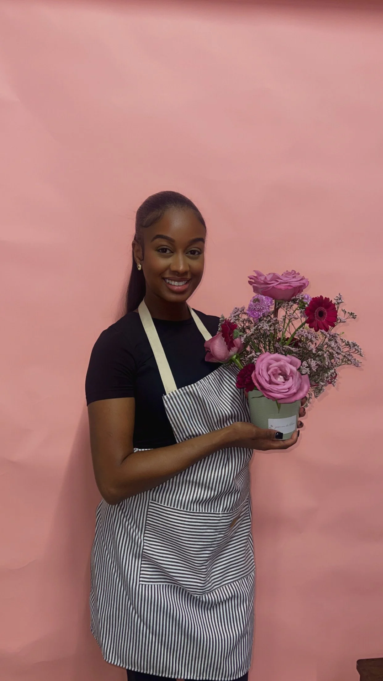 A smiling woman in a black shirt and a striped apron holding a pot of pink and purple flowers, standing against a pink background.