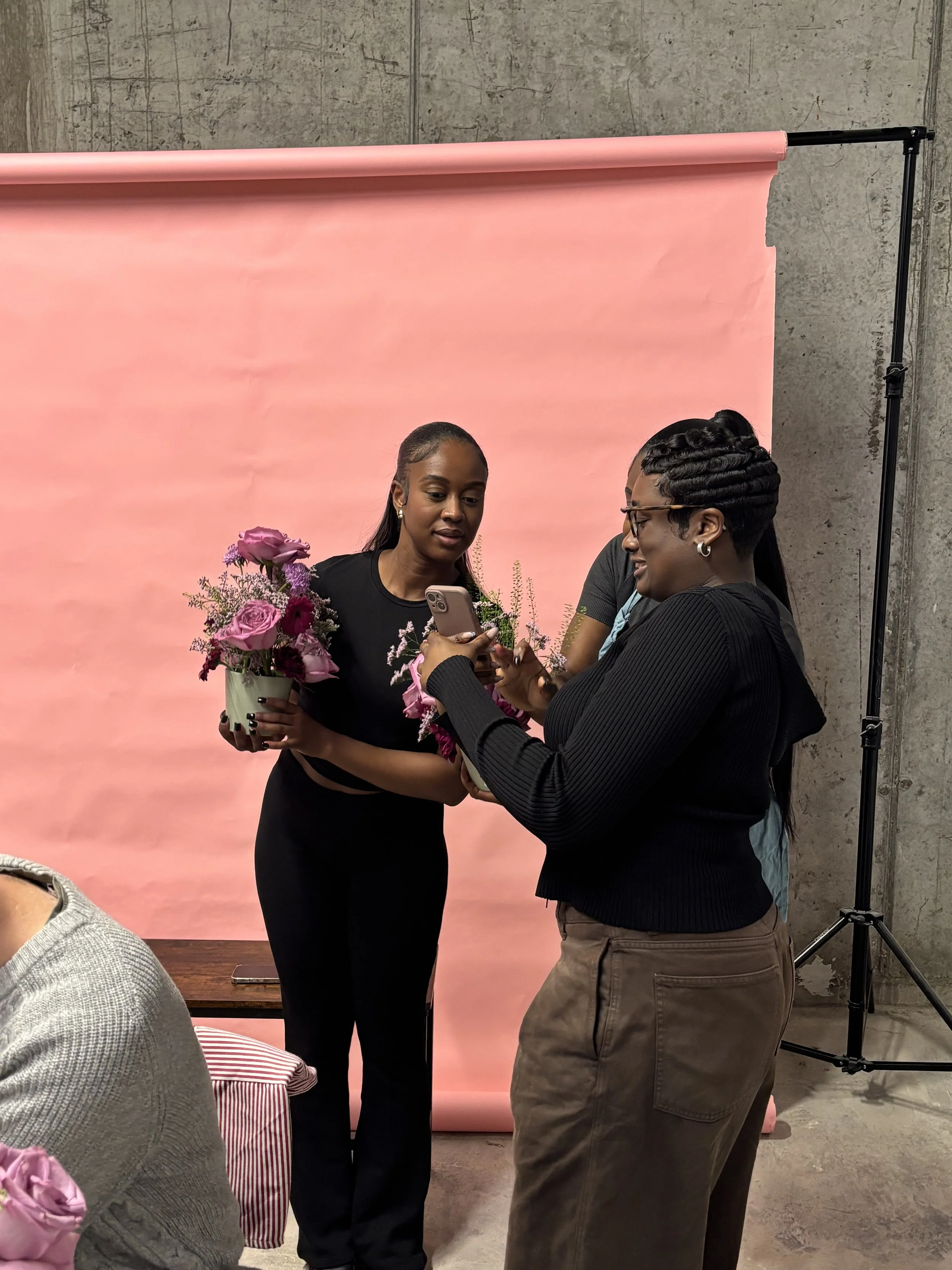 Two women at a photoshoot with a pink backdrop; one woman holds a bouquet of pink flowers, and the other woman shows her something on her phone.