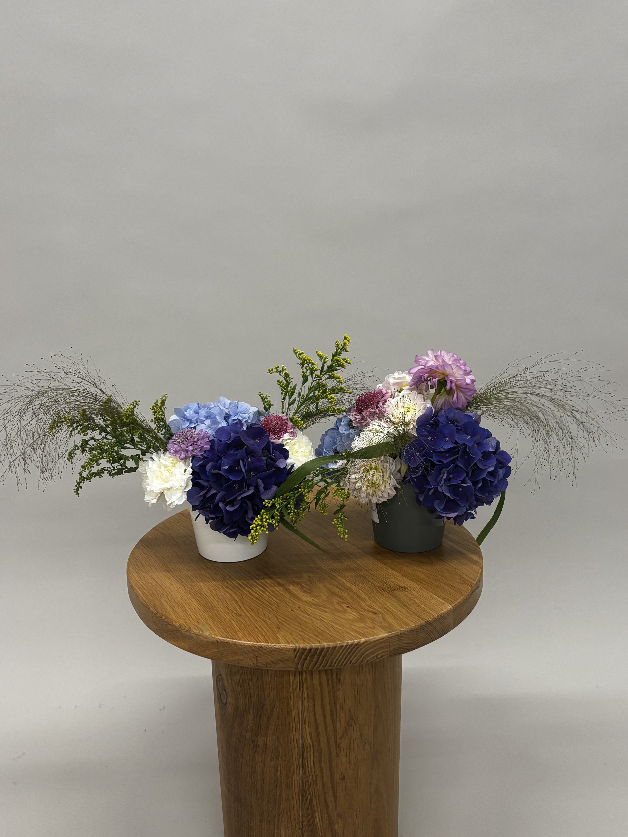 Two floral arrangements with purple, blue, white, and pink flowers and greenery on a round wooden pedestal table against a plain light-colored background.