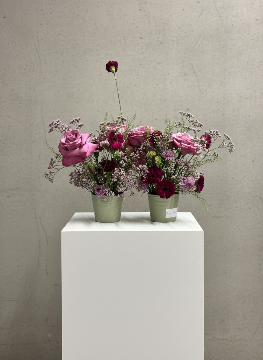 Two arrangements of pink and purple flowers in green pots placed on a white pedestal against a plain beige wall.