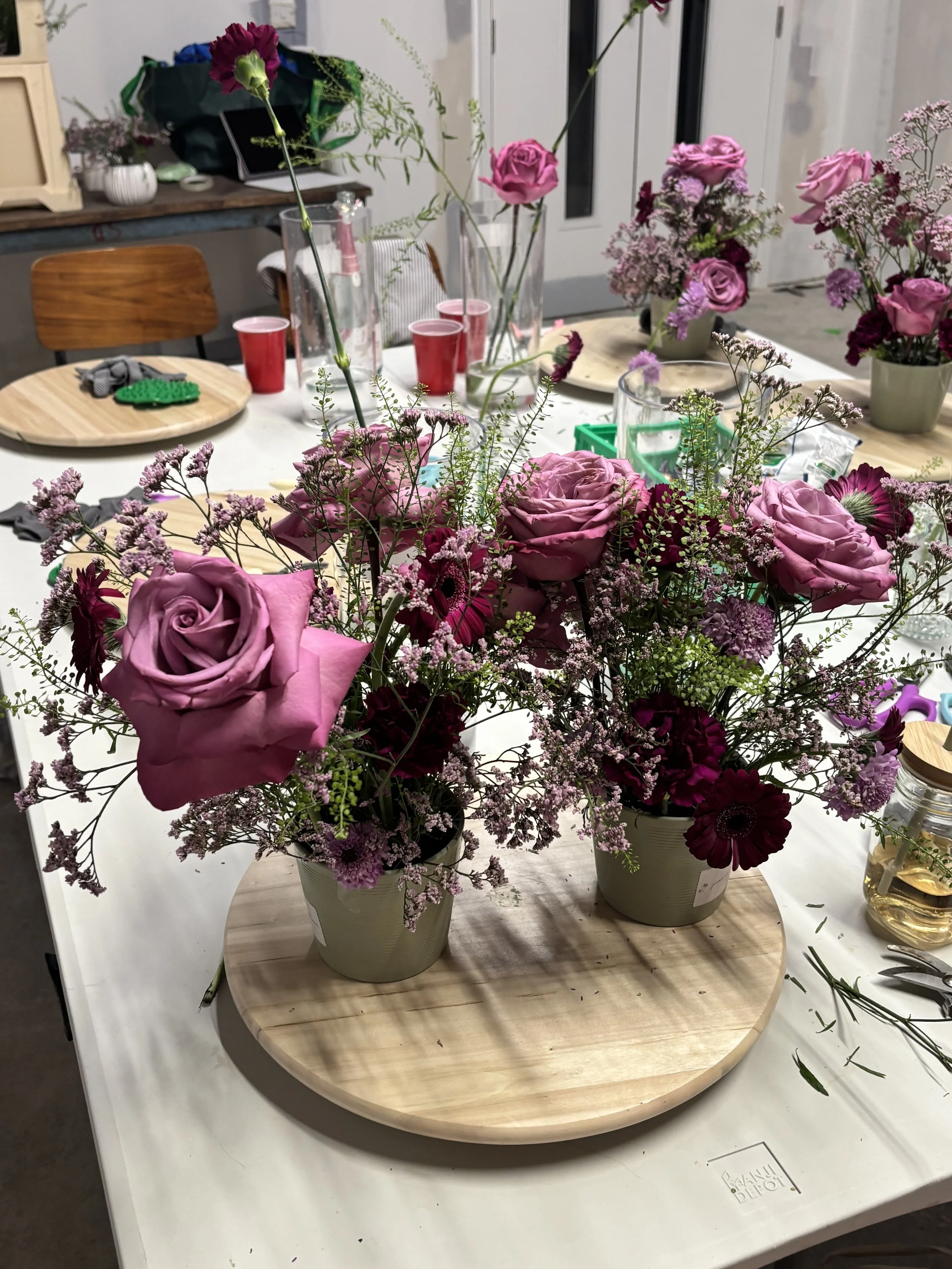 Several pink and purple flower arrangements in small green pots on a light wooden tray, set on a white table. The background has additional flowers, vases, cups, and tools for flower arranging.