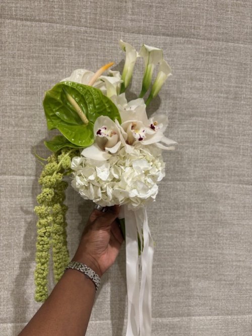 Hand holding a white floral bouquet with calla lilies, orchids, hydrangeas, and greenery, tied with a white ribbon, against a textured beige background.