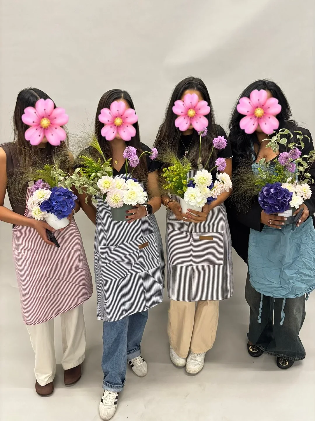 Four women with face flowers holding flower arrangements in vases, standing against a plain wall.