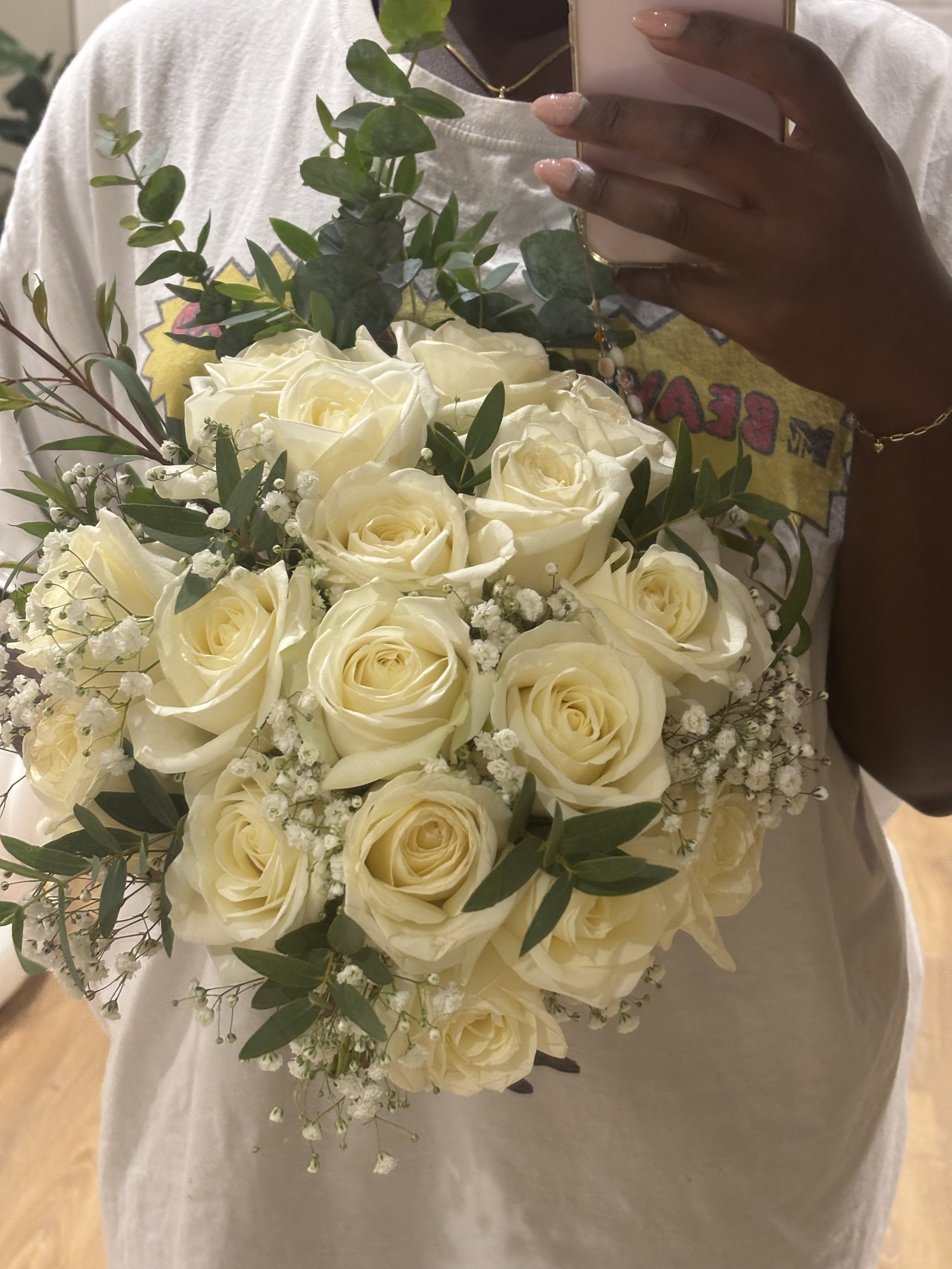 Person holding a smartphone with a white and cream flower bouquet, including roses and baby's breath, in front of them.