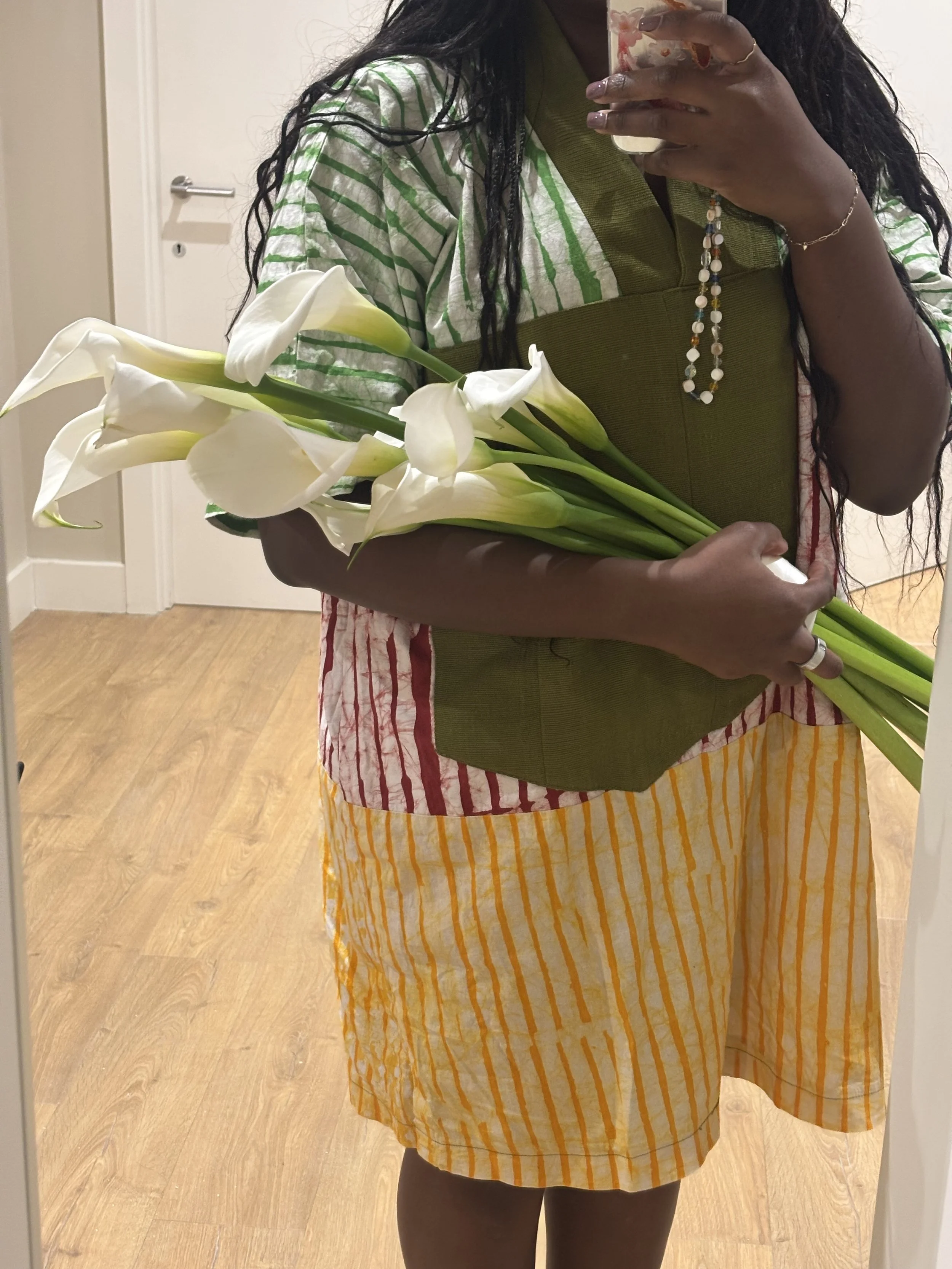 Person taking a mirror selfie holding a bouquet of white calla lilies, wearing a multicolored striped dress with green, red, and yellow hues, and a layered necklace.
