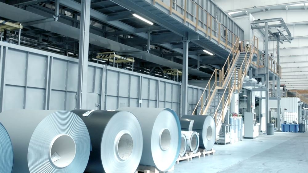Interior of a manufacturing plant with large rolls of Zinc sheet metal, industrial equipment, and a staircase leading to an upper level.