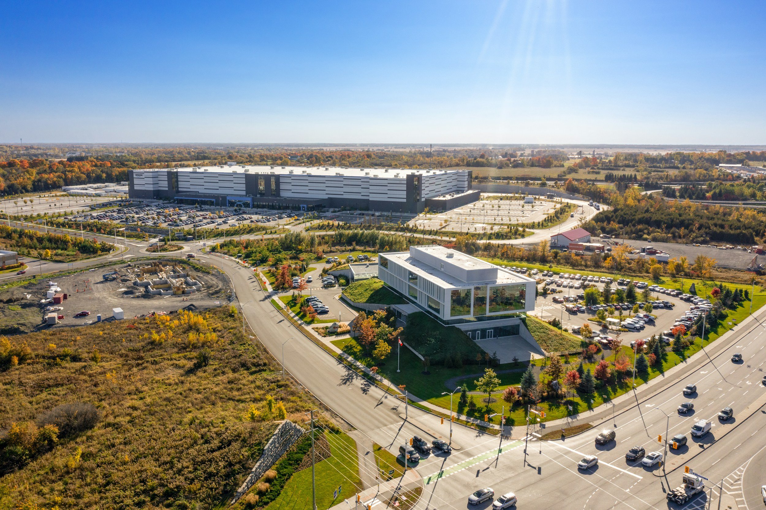 Aerial view of a large modern building and parking lots surrounded by trees with fall foliage, under a clear blue sky.