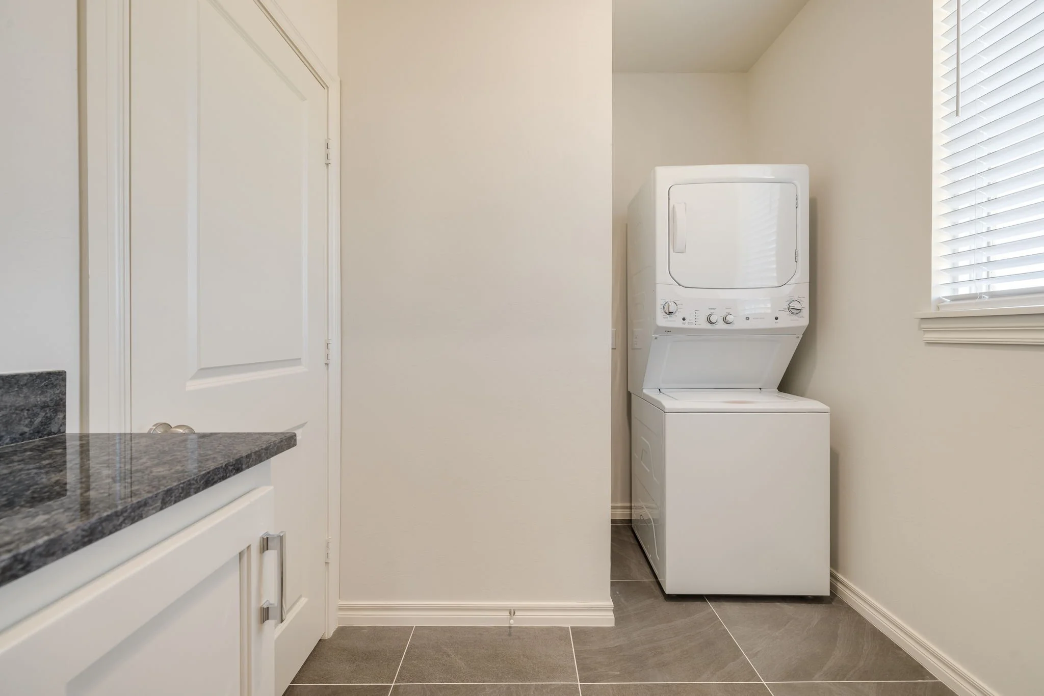 A laundry room with a window, a white stacked washer and dryer, white cabinets, and tiled flooring.