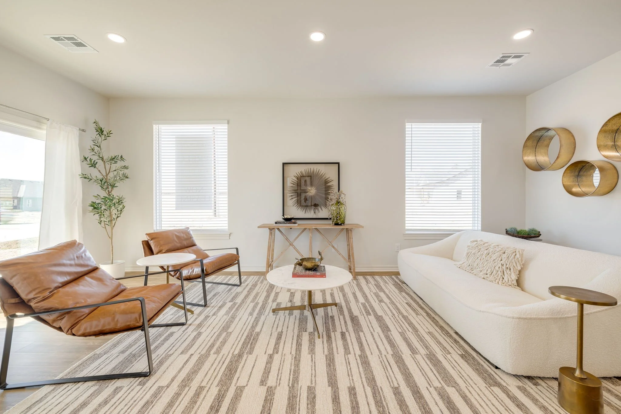 A modern living room with white walls, large windows with blinds, and ceiling lights. There's a white sofa with a plush pillow, a round marble-top coffee table, and two brown leather lounge chairs. Decorative items include wall art, gold circular shelves, and a potted plant.