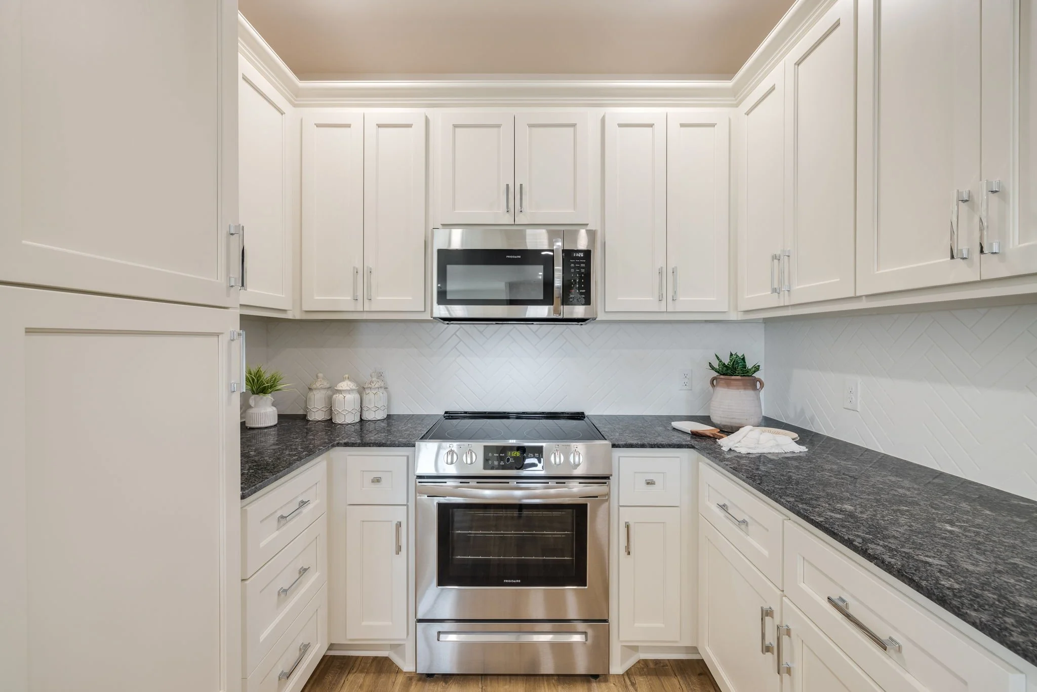 White kitchen with granite countertops, stainless steel microwave above oven, and decorative jars and plants on countertop.