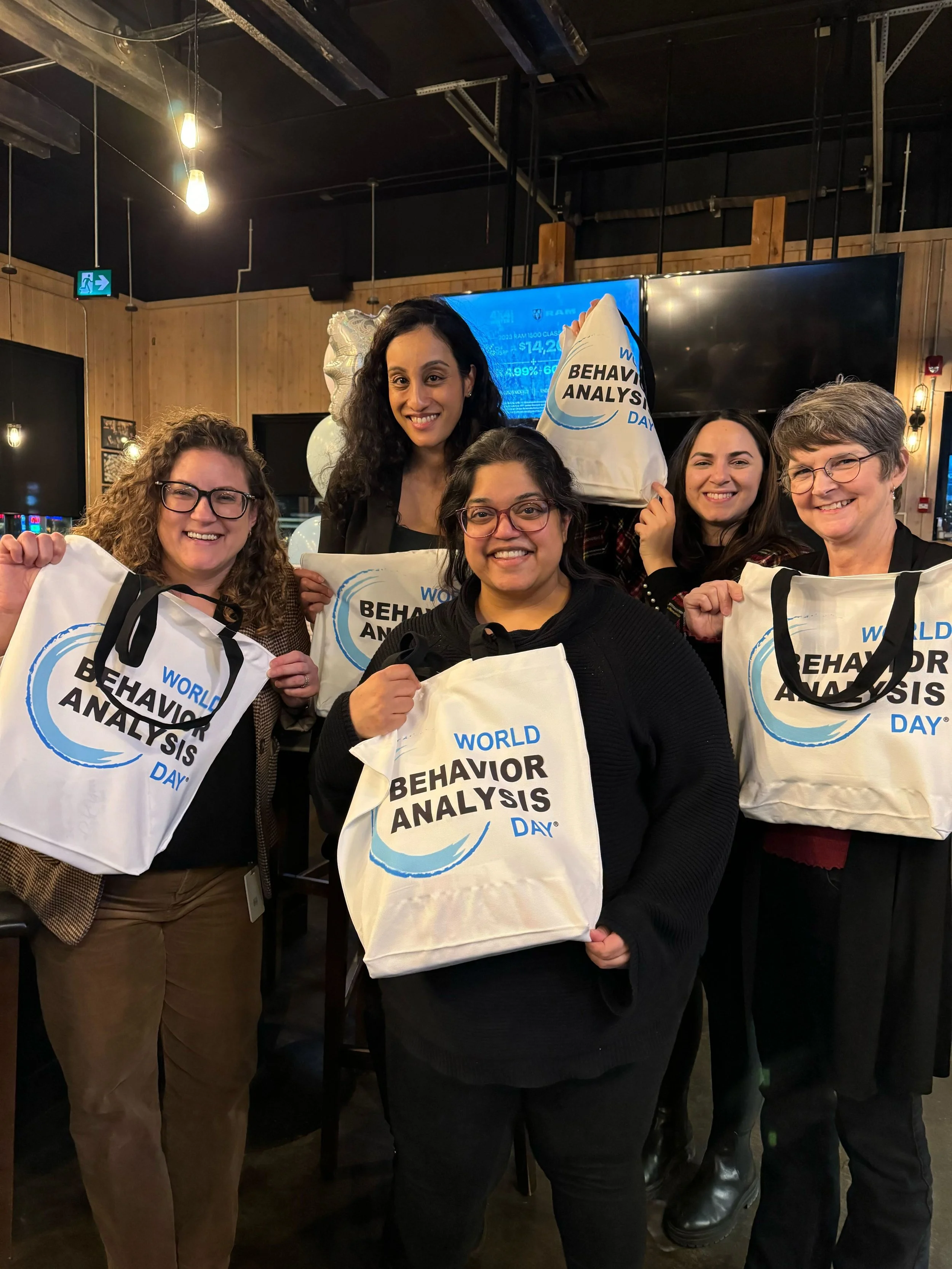 A photograph of five smiling women standing together in a casual event space with warm pendant lighting. They are proudly holding up white canvas tote bags that feature the text "WORLD BEHAVIOR ANALYSIS DAY" alongside a blue swoosh graphic.