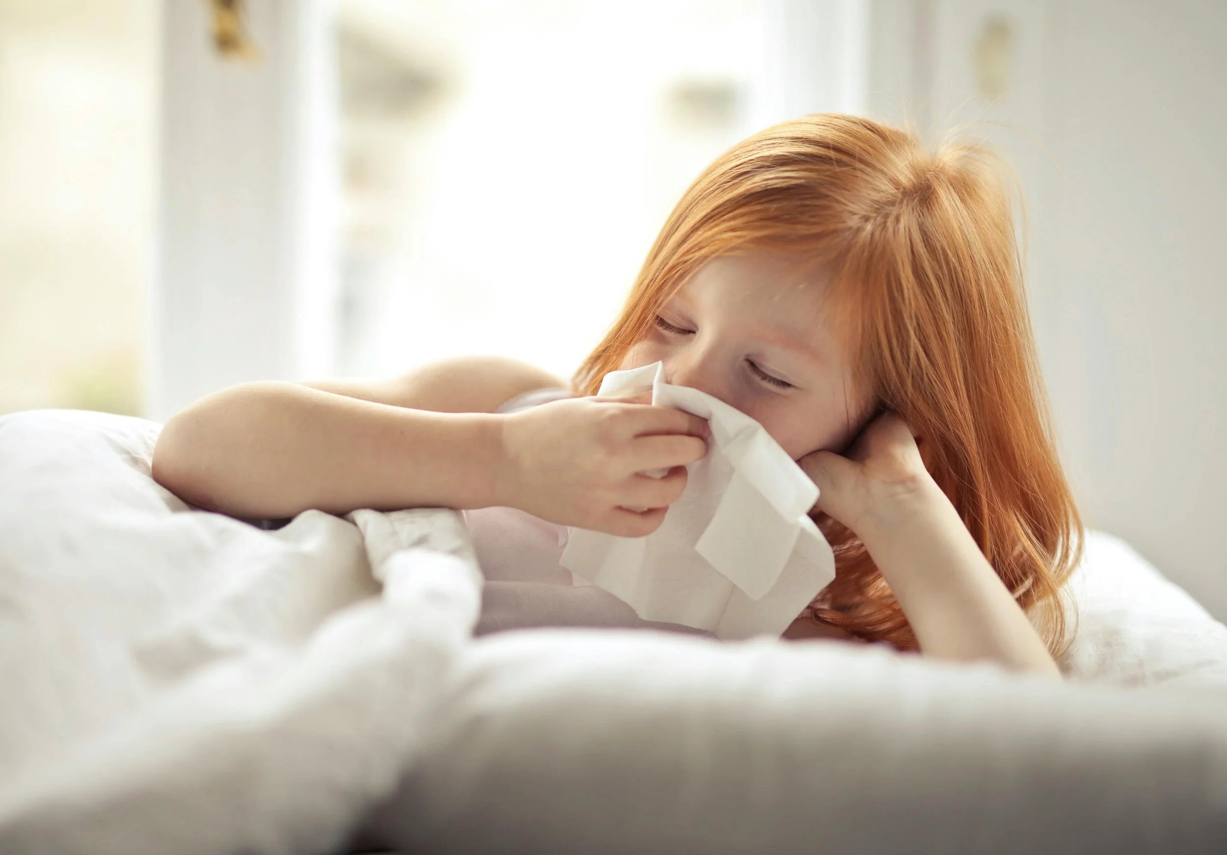 A child with red hair lying in bed and using a tissue to blow their nose.