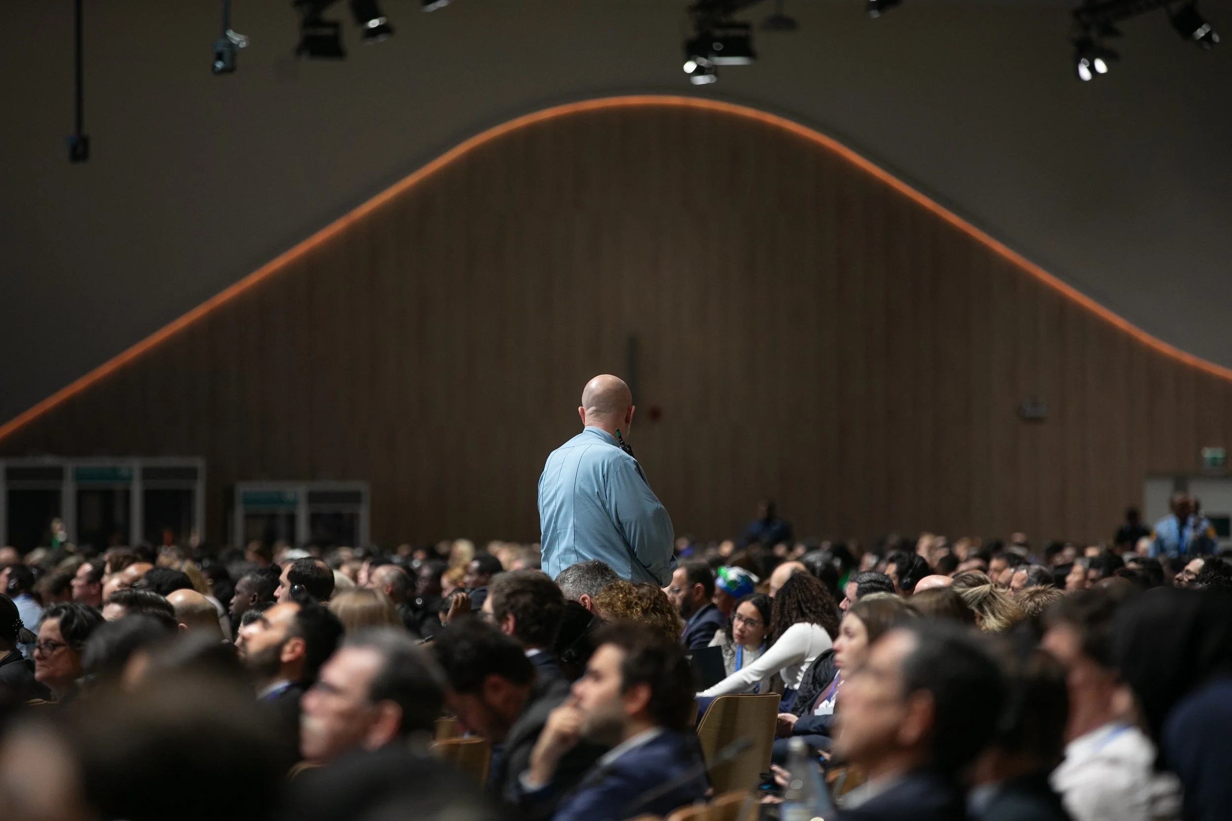 A man in a light blue shirt stands in front of a large audience in a conference hall, with a high ceiling and wood-paneled walls - www.brentkerrigan.com - photo by Kiara Worth