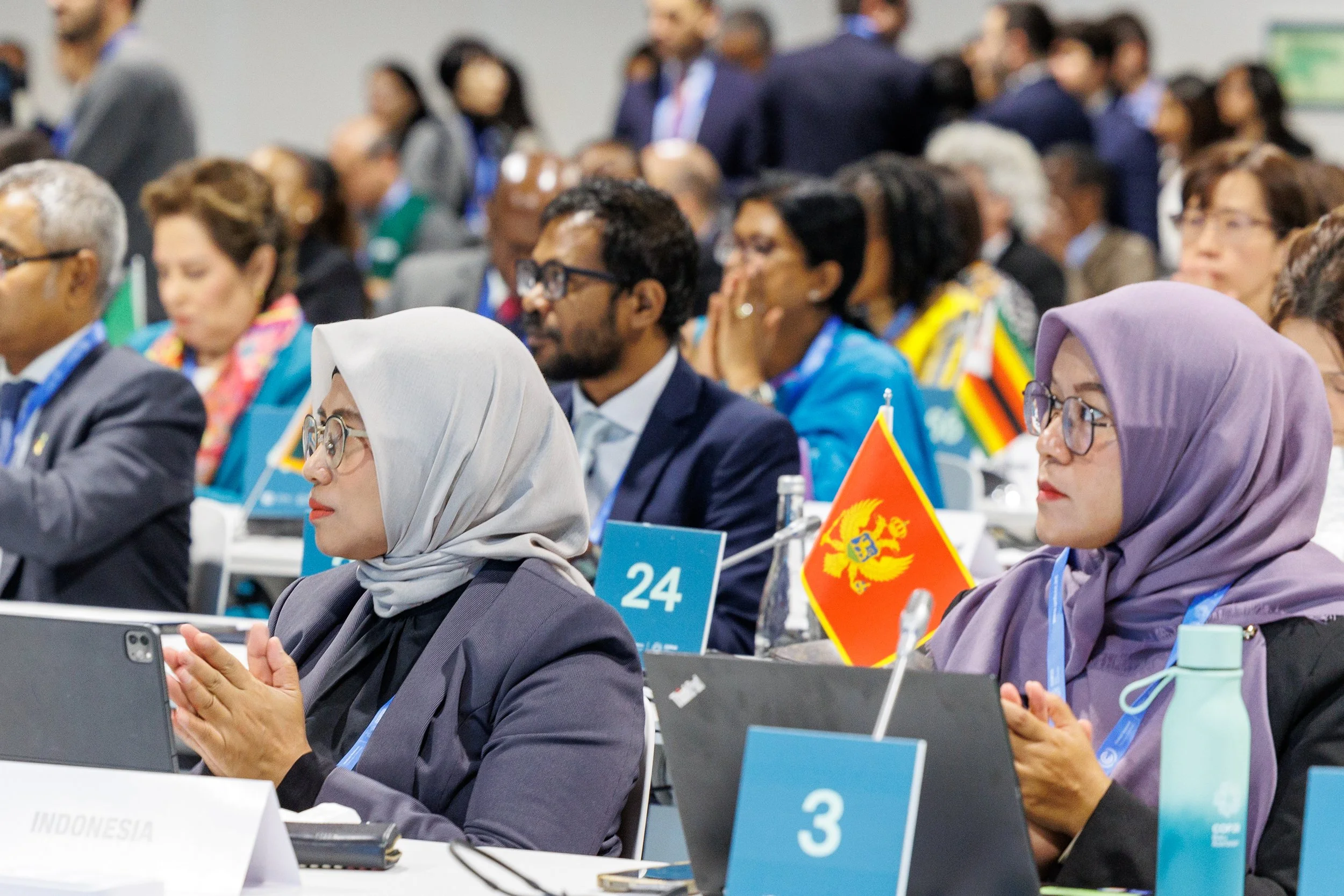 Group of diverse women and men attending a conference, some wearing headscarves, glasses, and business attire, sitting at tables with flags and conference materials - www.brentkerrigan.com - photo by Kiara Worth
