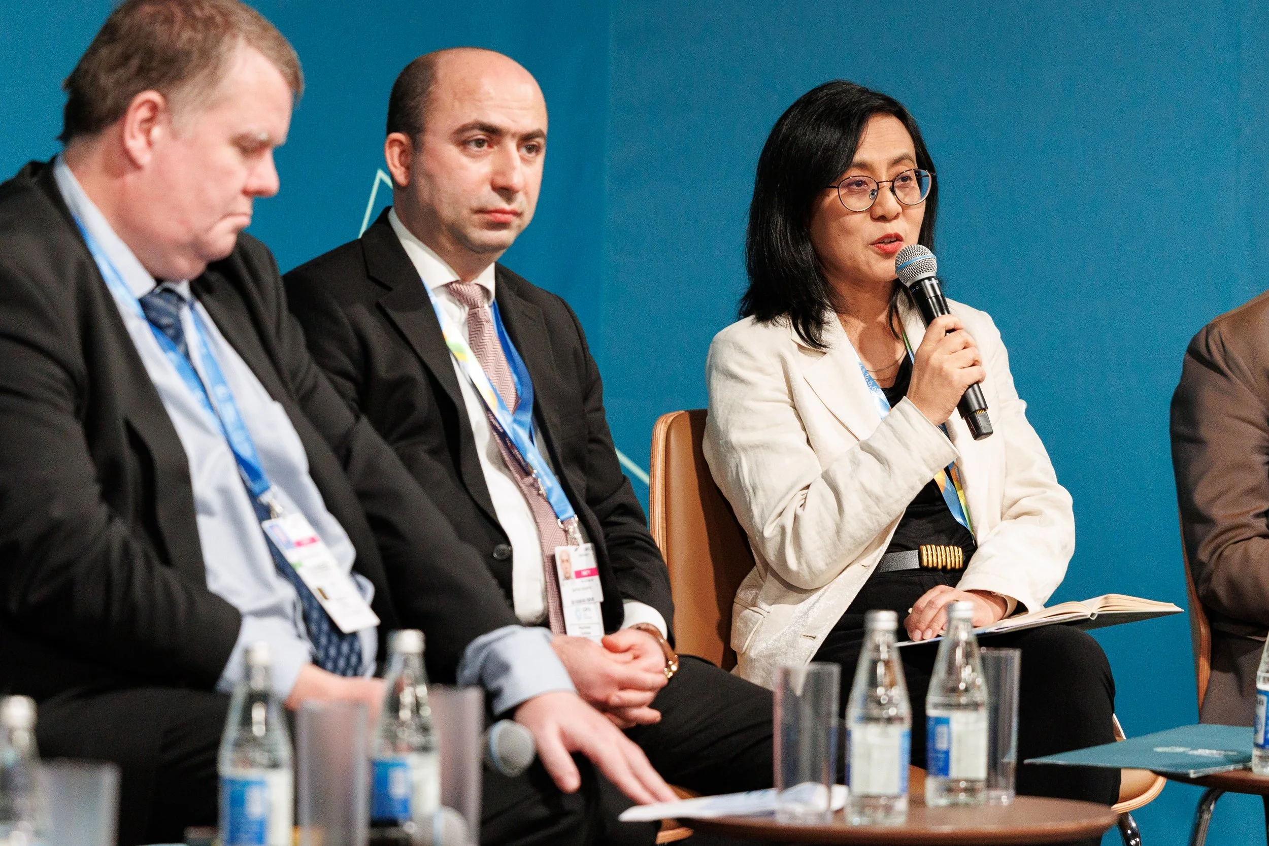 Four people sit at a panel discussion, with the woman on the far right holding a microphone, against a blue background. Bottles of water are on the table in front of them - www.brentkerrigan.com - photo by Kiara Worth
