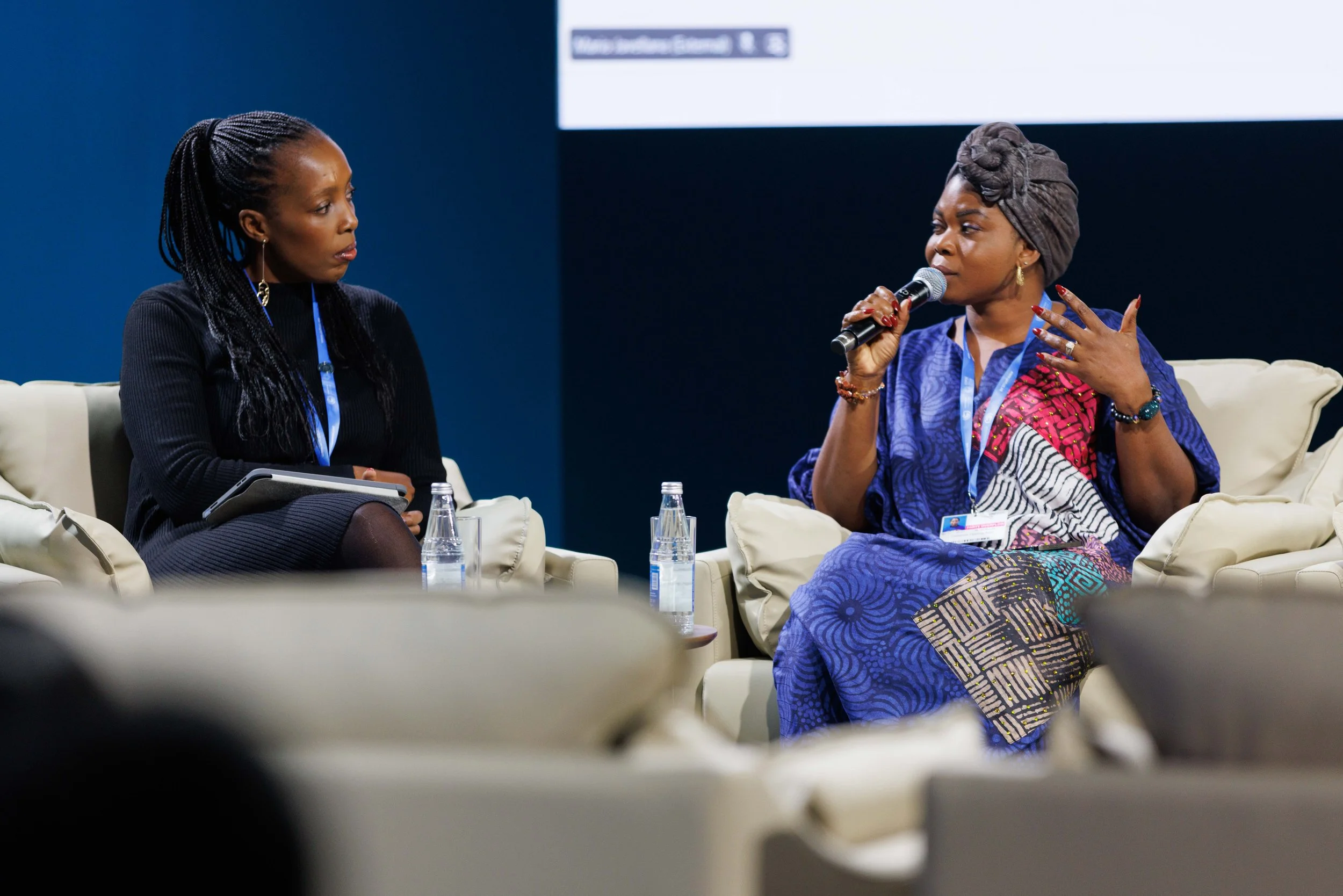 Two women sitting on a stage engaged in conversation during a panel discussion. One woman is speaking into a microphone, while the other woman listens attentively - www.brentkerrigan.com - photo by Kiara Worth