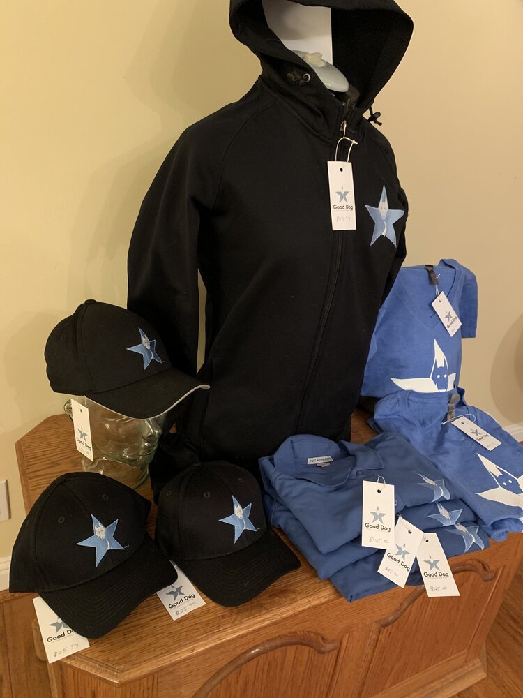 Black and blue hats and hoodies with a federally trademarked, star logo and tags that say "Good Dog," displayed on a wooden table. Dog training apparel for small business for dog training.