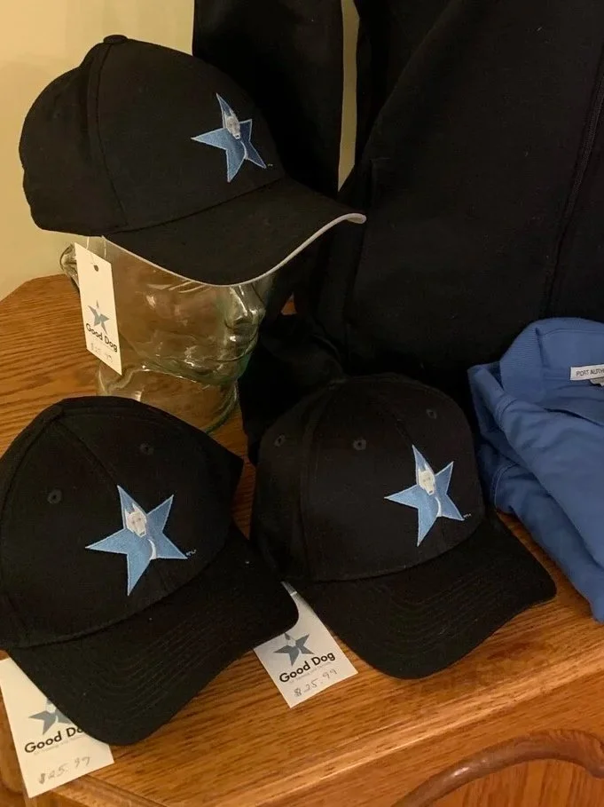 Black baseball caps with a blue star logo on top of a wooden table, with tags that read 'Good Dog' and a price of $25.99, and a mannequin head wearing one of the caps.