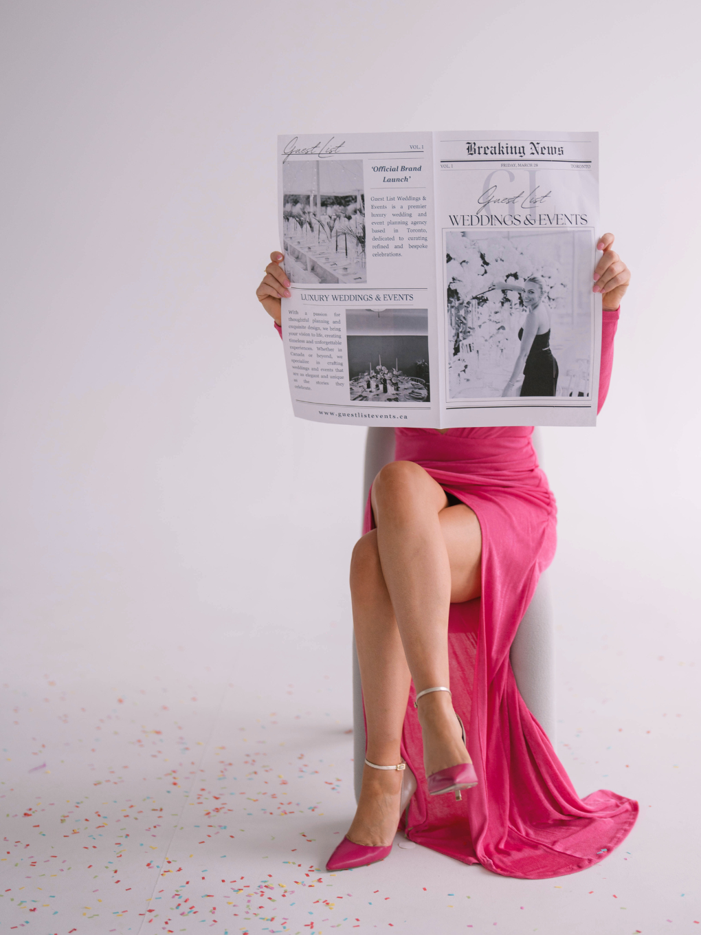 Person in a pink dress with crossed legs, wearing pink high heels, sitting on a chair, holding a newspaper in front of their face, with colorful confetti on the floor.