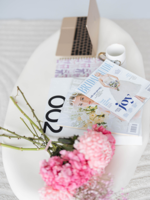 A white bathtub holding a bouquet of pink flowers, a magazine, a notebook, a pair of glasses, a mug, and a laptop.