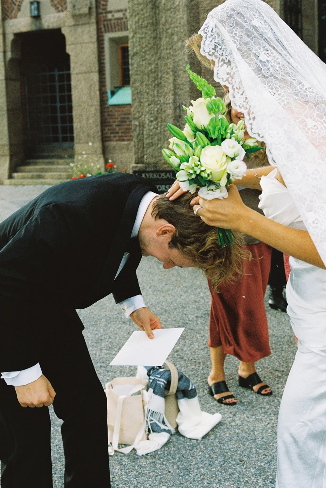 Bride in a white lace dress holding a bouquet of white and green flowers, performing a blessing or ritual on groom in a black suit, who is bowing his head and holding a note, outside on a street with stairs and building in the background.