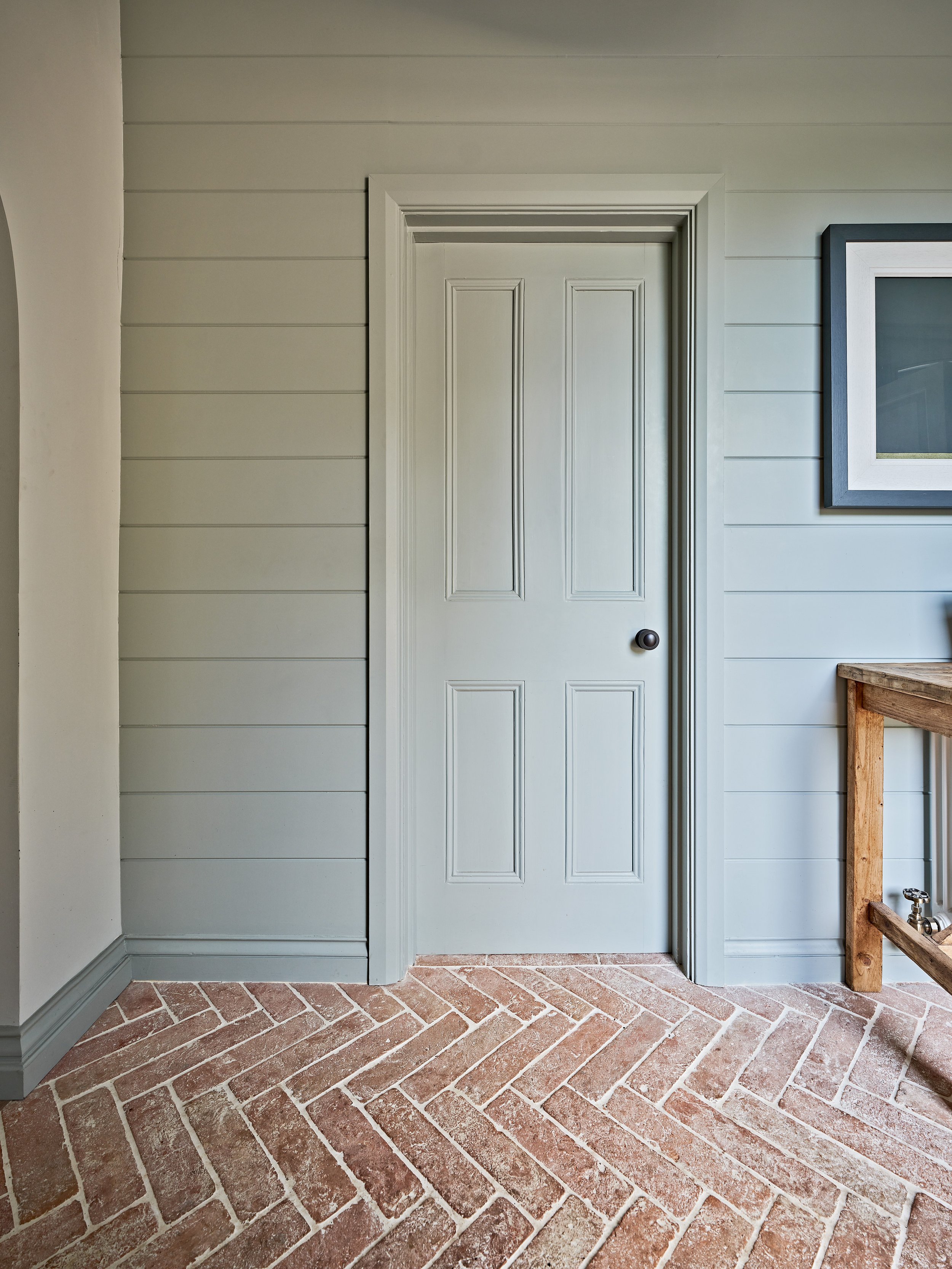 Interior room with a light blue paneled door, wooden wall paneling, brick herringbone floor, and part of a framed picture.