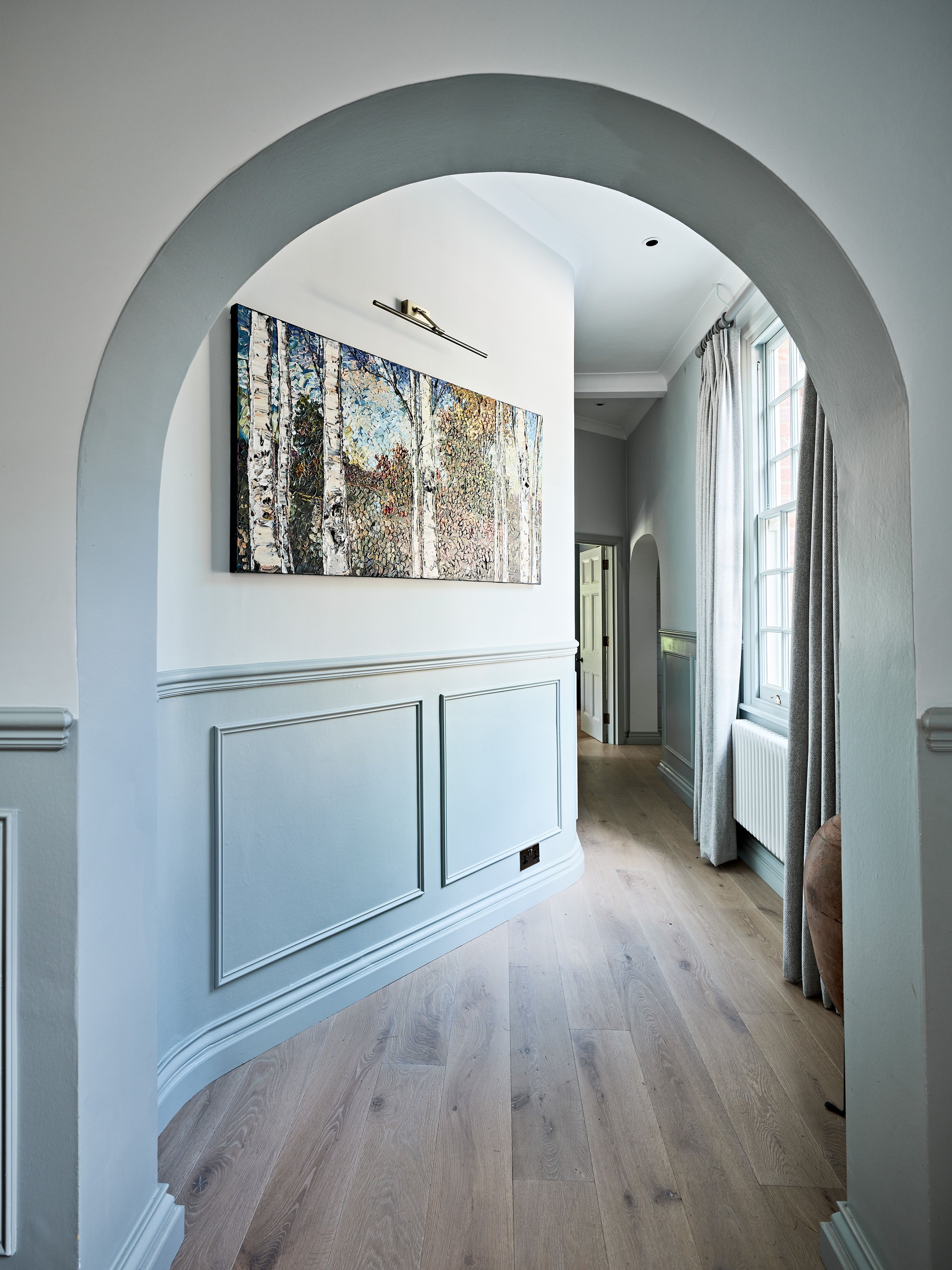 Elegant hallway with arched doorway, light wooden flooring, wainscoting, and a large artwork depicting birch trees on the wall.