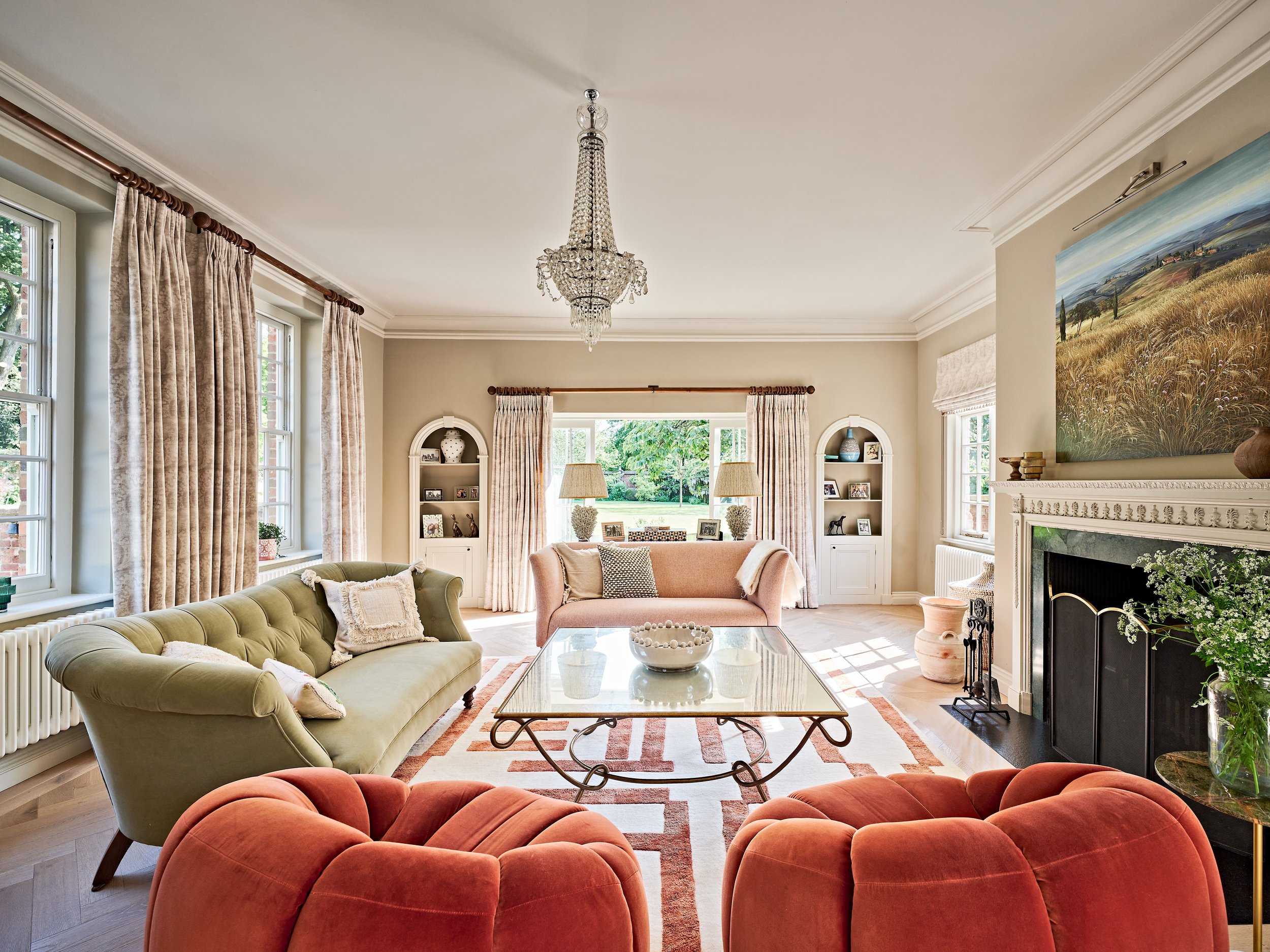 Elegant living room with a chandelier, sofas, and glass coffee table. Features large windows with curtains, a decorative fireplace, and built-in shelves with ornaments. Bright and airy atmosphere with natural light.