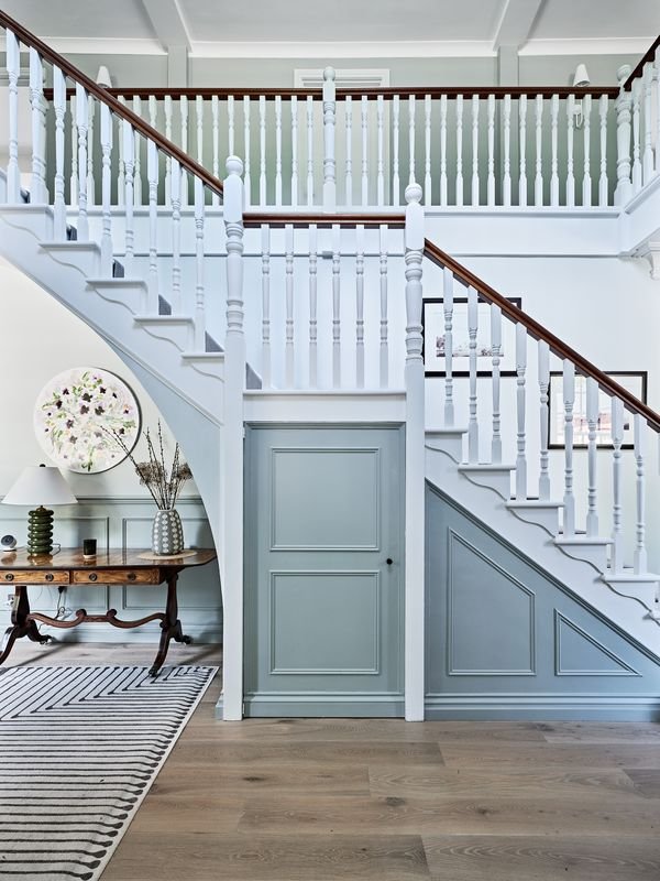 Interior of a house with a white staircase featuring wooden handrails and a light blue under-stair storage door. A striped carpet partially visible on wooden flooring. A wooden console table with decorative items, including a lamp, vase, and art, sit
