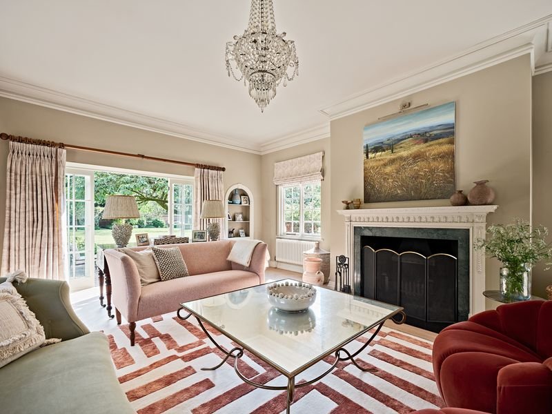 Elegant living room with chandelier, pink sofa, armchair, and glass coffee table. Features a fireplace with artwork above, striped rug, large window with curtains, and decorative shelves.