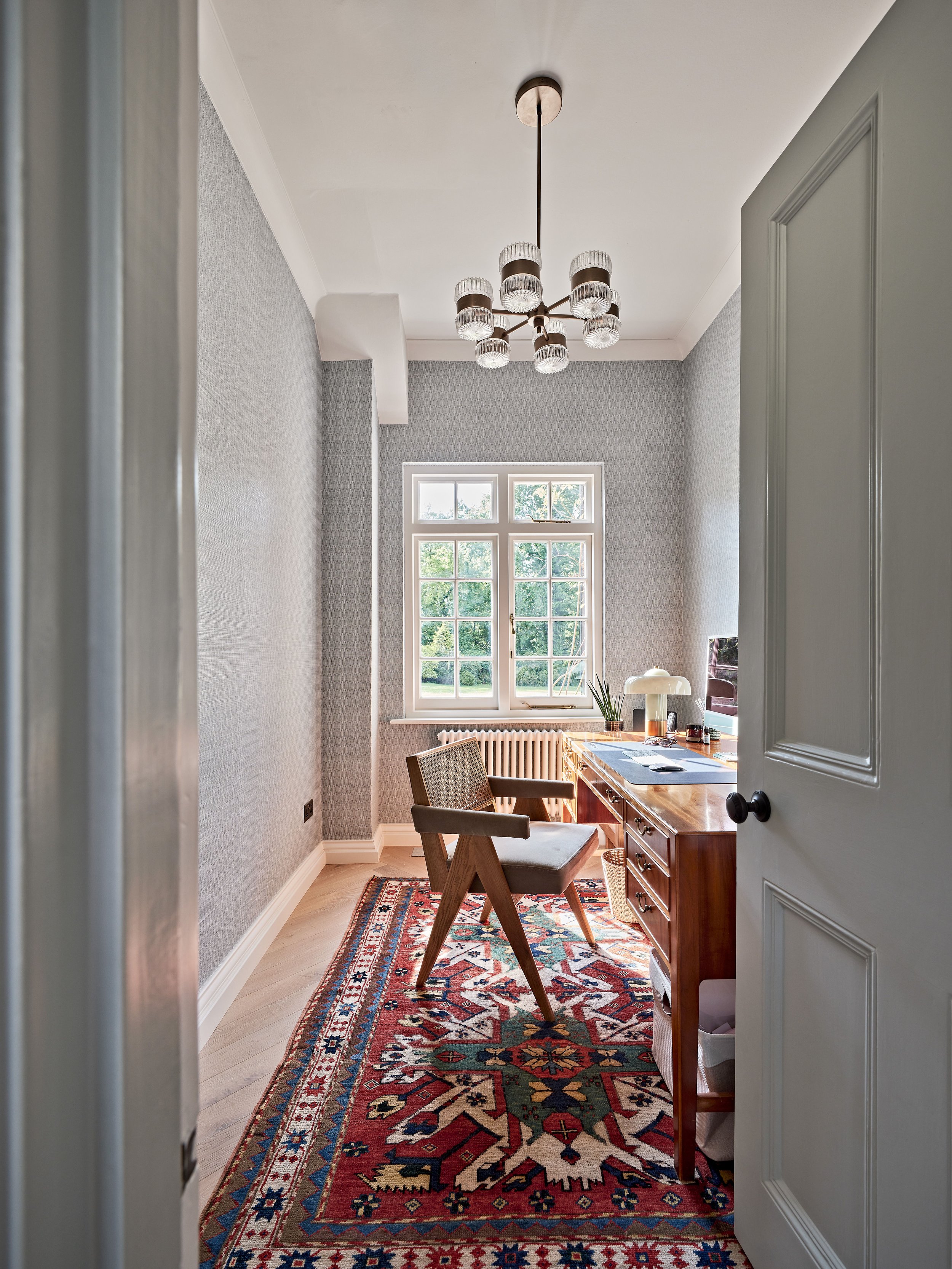 A small home office with a patterned red area rug and wooden desk. The room has a large window with natural light, a woven chair, and a ceiling light fixture above. The walls are light-colored and the flooring is wooden.