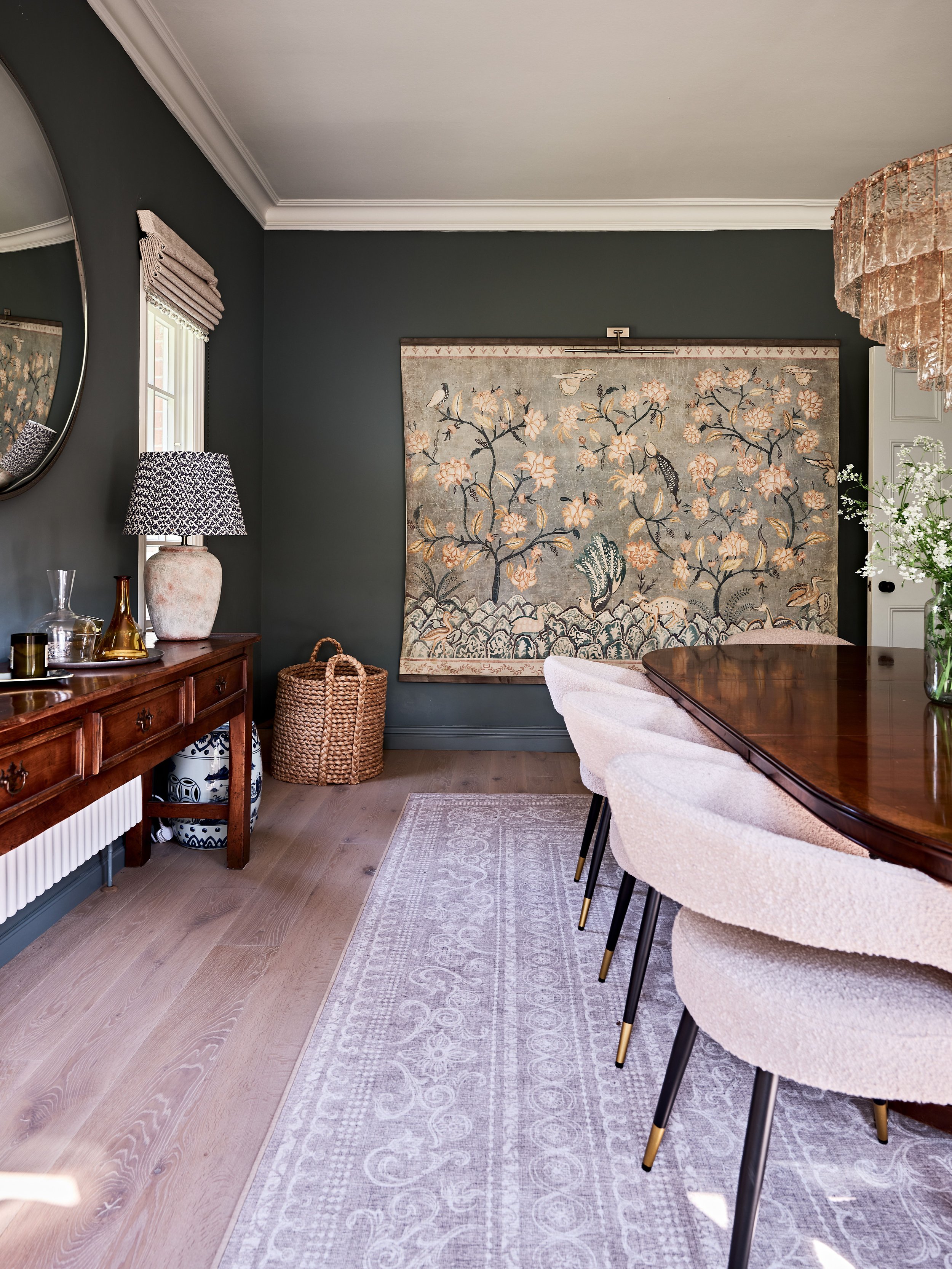 Elegant dining room with dark walls, featuring a wooden table and upholstered chairs, a large wall tapestry with floral motifs, a wooden sideboard with decorative objects, and a woven basket on the floor.