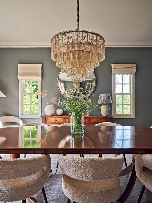 A dining room with a wooden table and cream-colored upholstered chairs. A large glass vase with white flowers is in the center of the table. The room features a vintage chandelier, two windows with beige Roman shades, a round mirror, a wooden sideboa