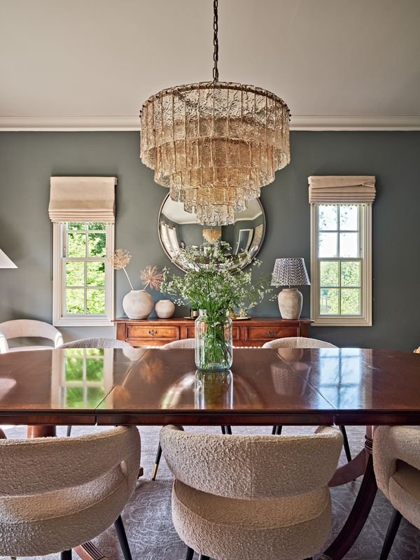 Elegant dining room with a large wooden table, surrounded by cushioned chairs, featuring a glass vase with flowers. A vintage chandelier hangs above. Background includes a sideboard, mirror, and windows with Roman shades.