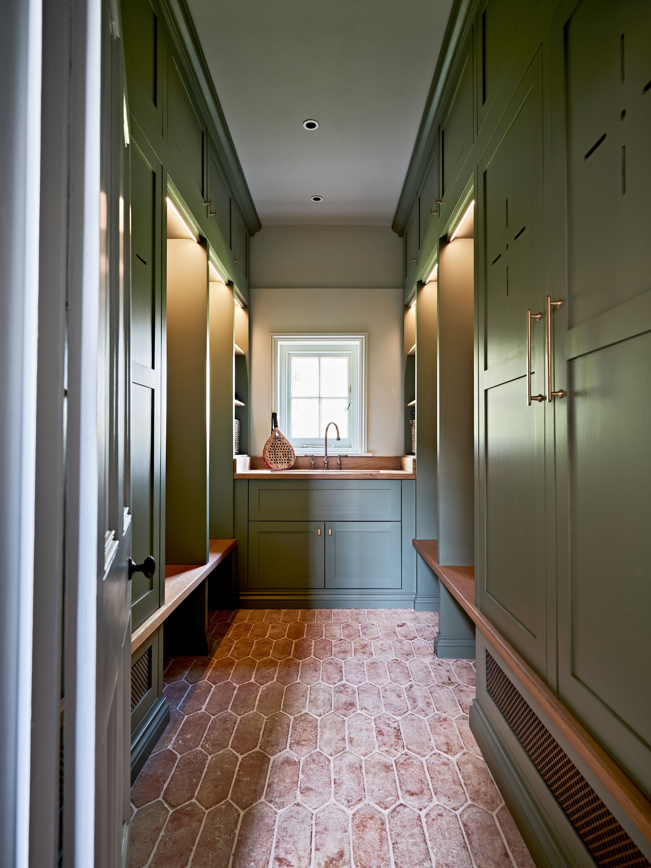 Modern green mudroom with built-in cabinets, hexagonal tile floor, wooden benches, and a small window above a sink with a decorative basket.
