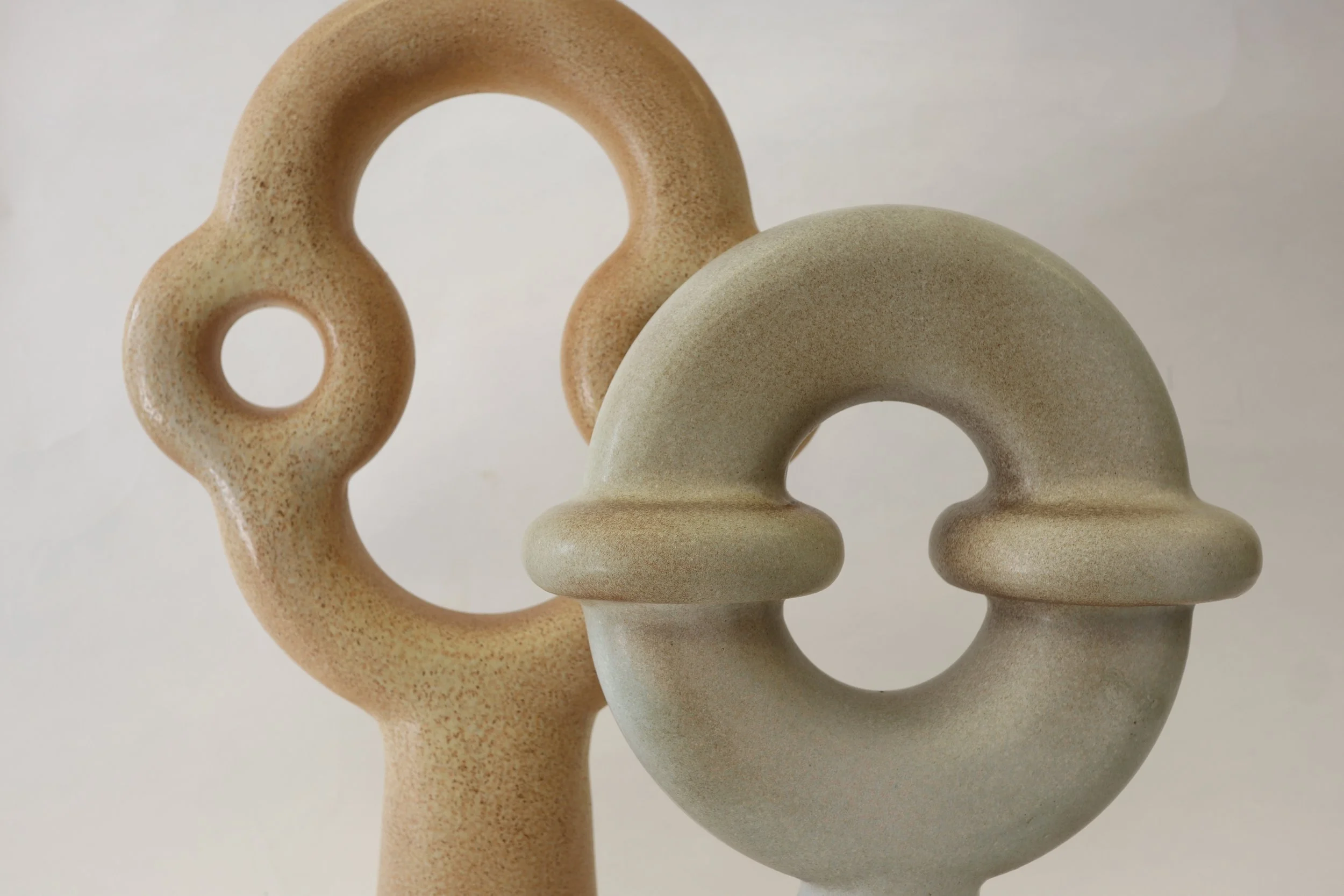 Two ceramic sculptures with abstract, rounded shapes and holes, in beige, tan, and light gray colors, against a plain white background.