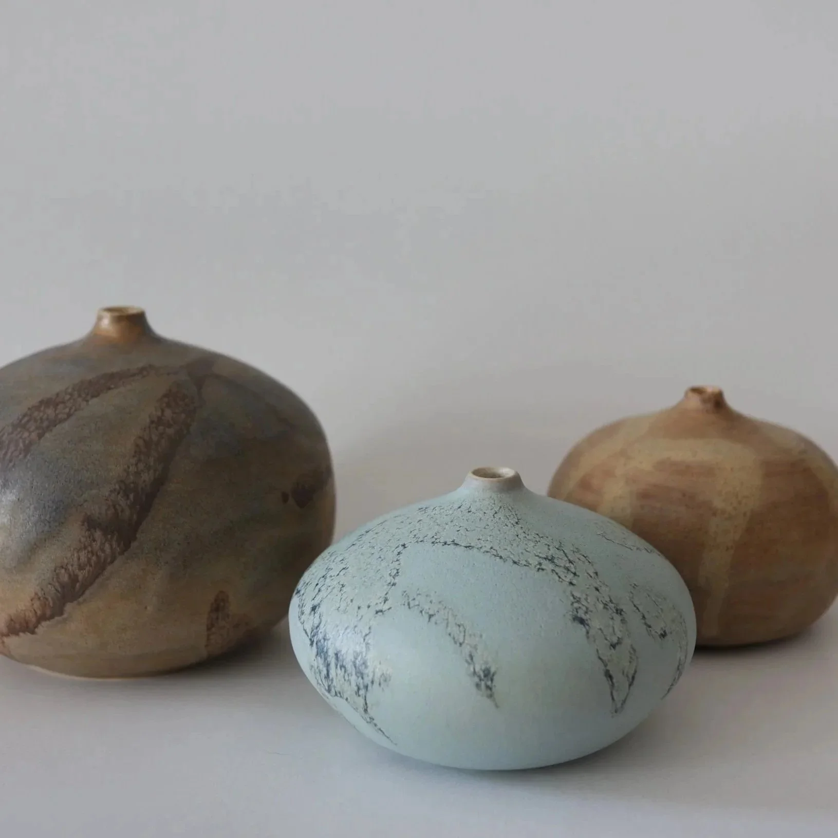 Three round ceramic vases with earth-tone and light blue glaze, arranged on a white surface against a plain light background.