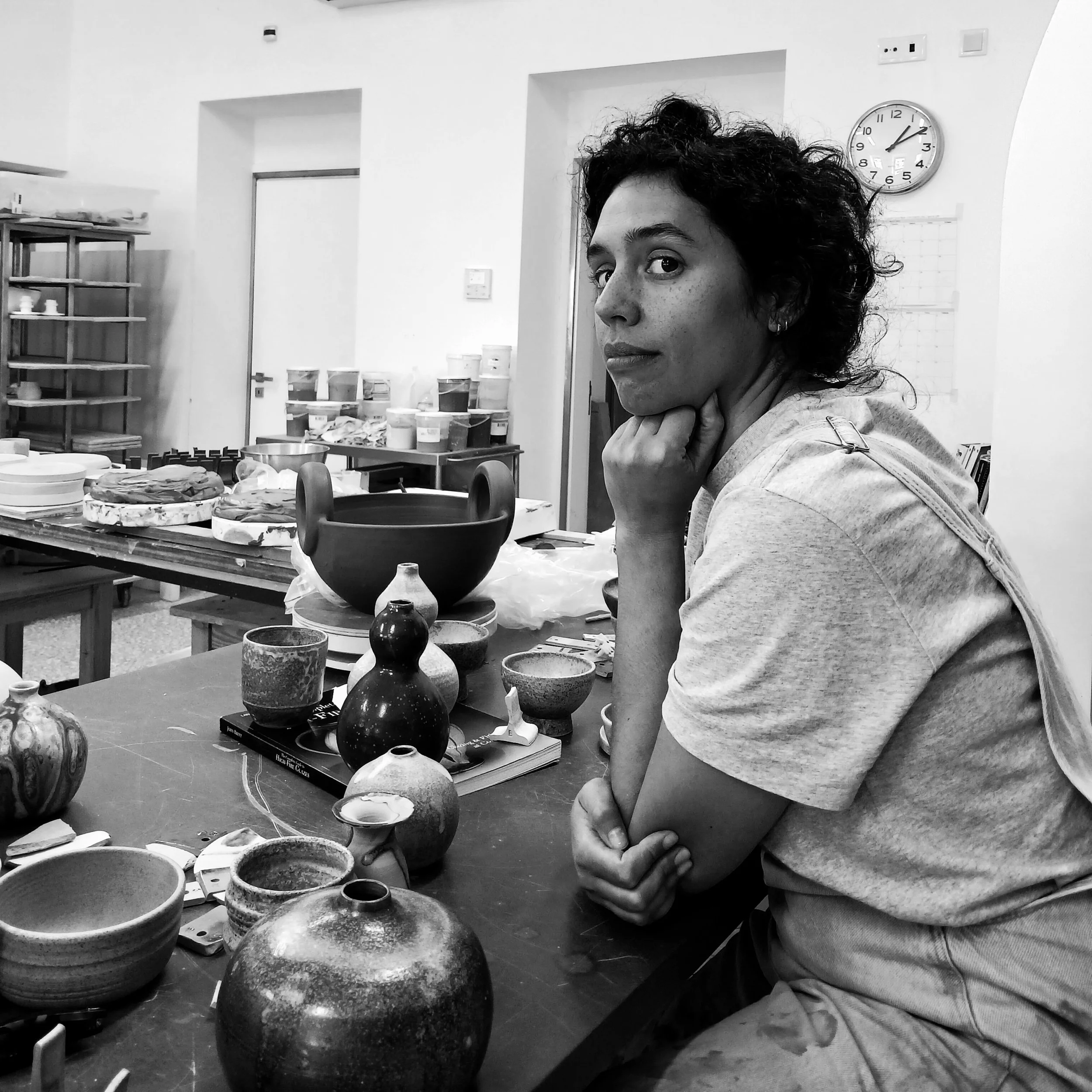 A woman with curly hair sits at a table with pottery and ceramics, in a studio or workshop with shelves and containers in the background.